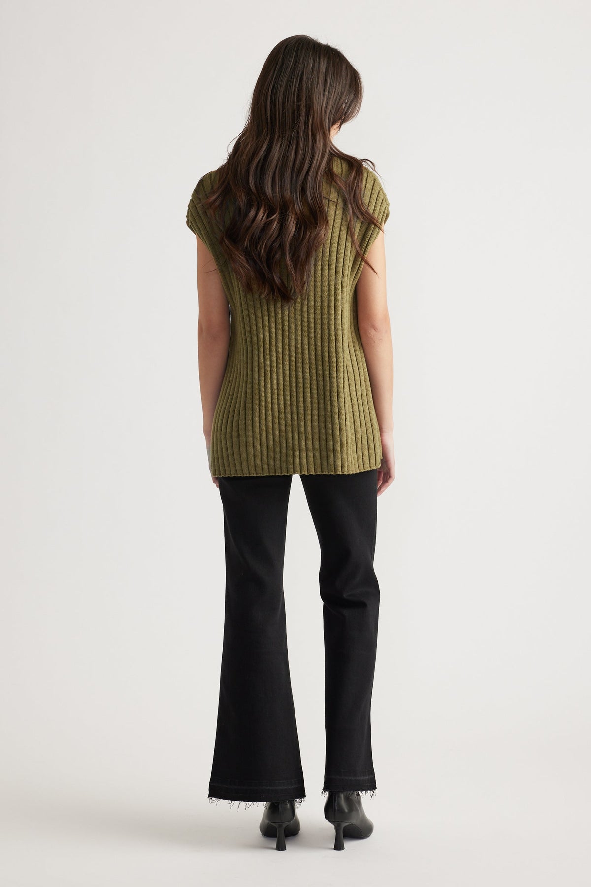 Woman with long wavy hair wearing an olive ribbed knit tunic paired with black flared jeans and heeled boots, back view.