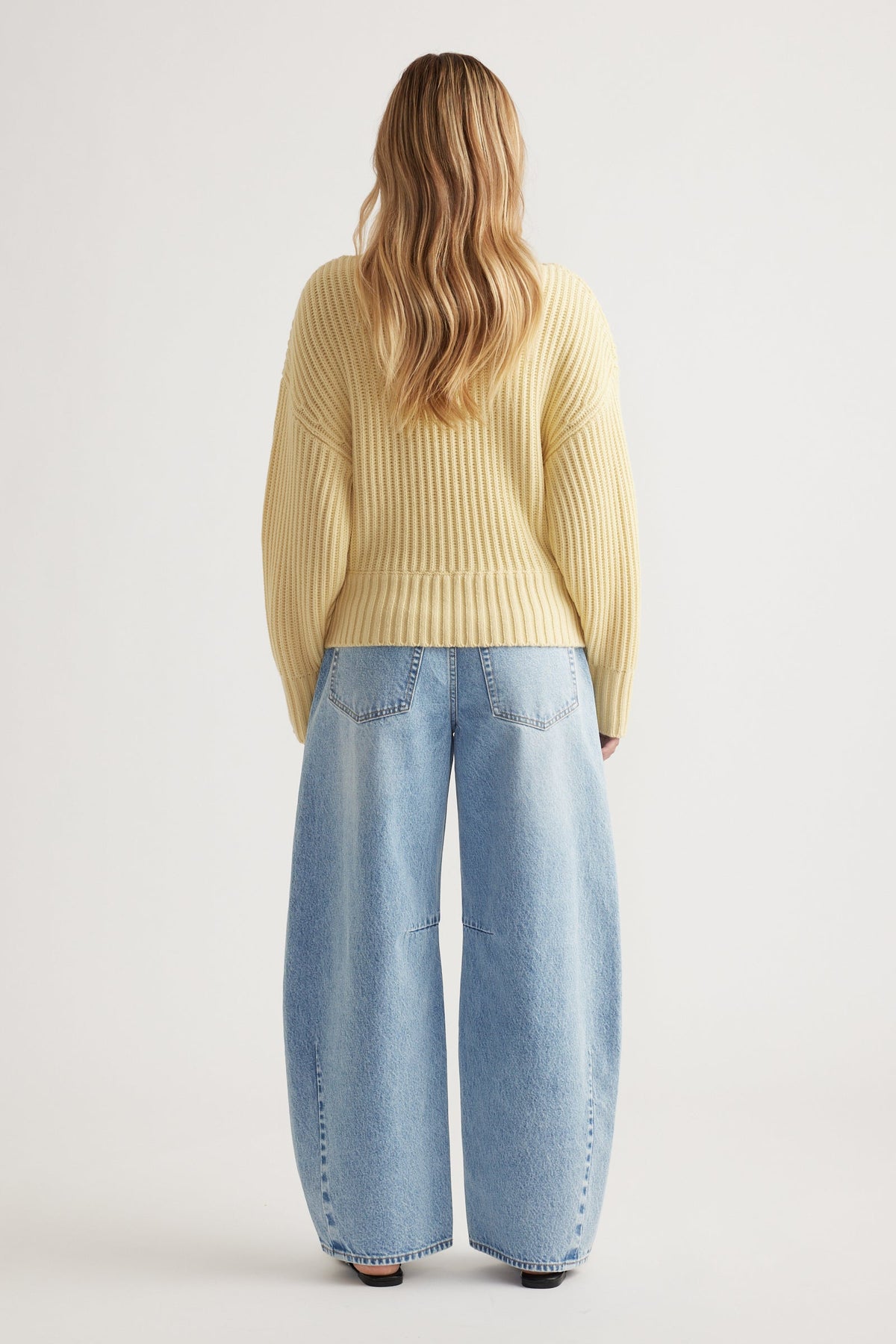 Back view of woman wearing a pale yellow drop shoulder knit jumper with relaxed wide-leg light denim jeans