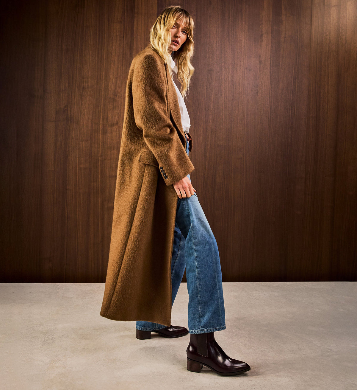 Person wearing a long brown coat, blue jeans, and brown chelsea boots against a wooden background