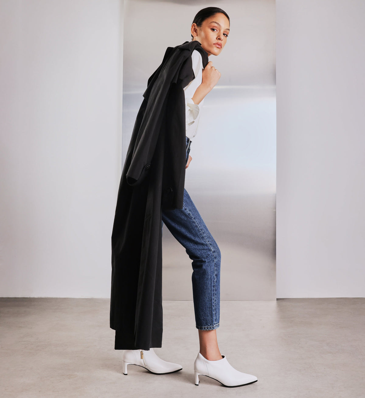 Woman wearing a black coat, white top, blue jeans, and white shoes in a minimalistic setting.