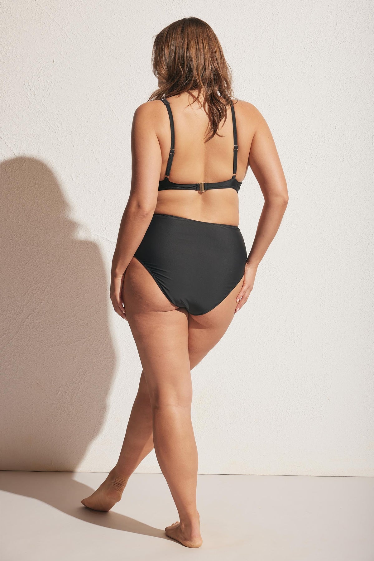 Woman wearing a black high-waisted bikini bottom and adjustable triangle bikini top, standing against a textured wall