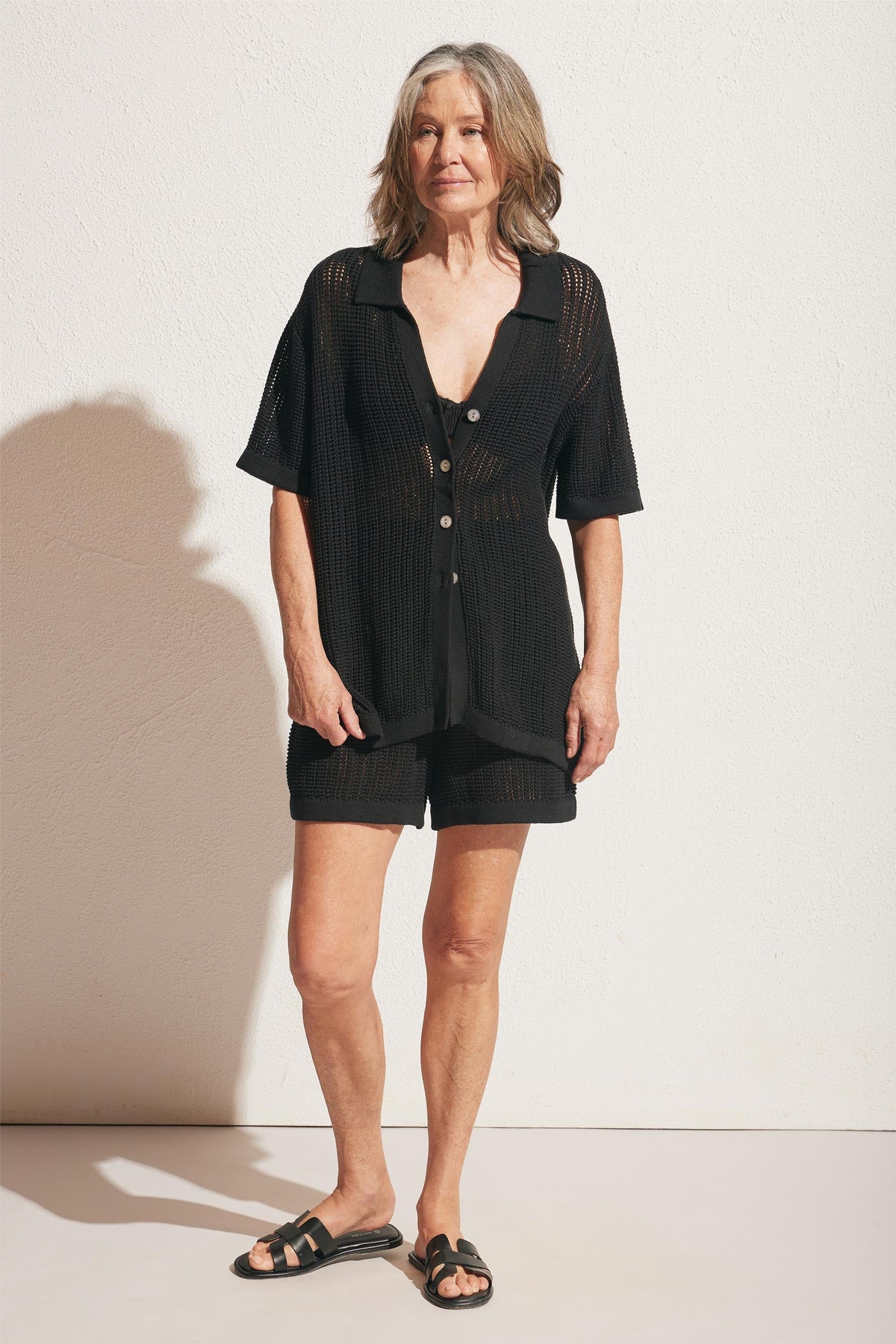 Woman wearing a black crochet knit short set with a button-up top and open-toe slide sandals, standing against a white wall.