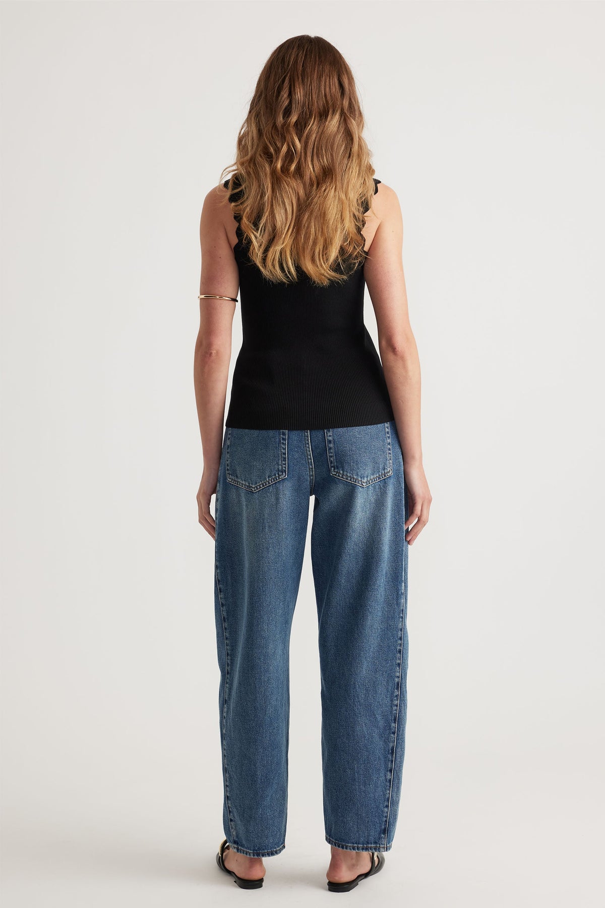 Woman wearing a fitted black tank with scallop edges paired with relaxed blue denim jeans, viewed from behind.