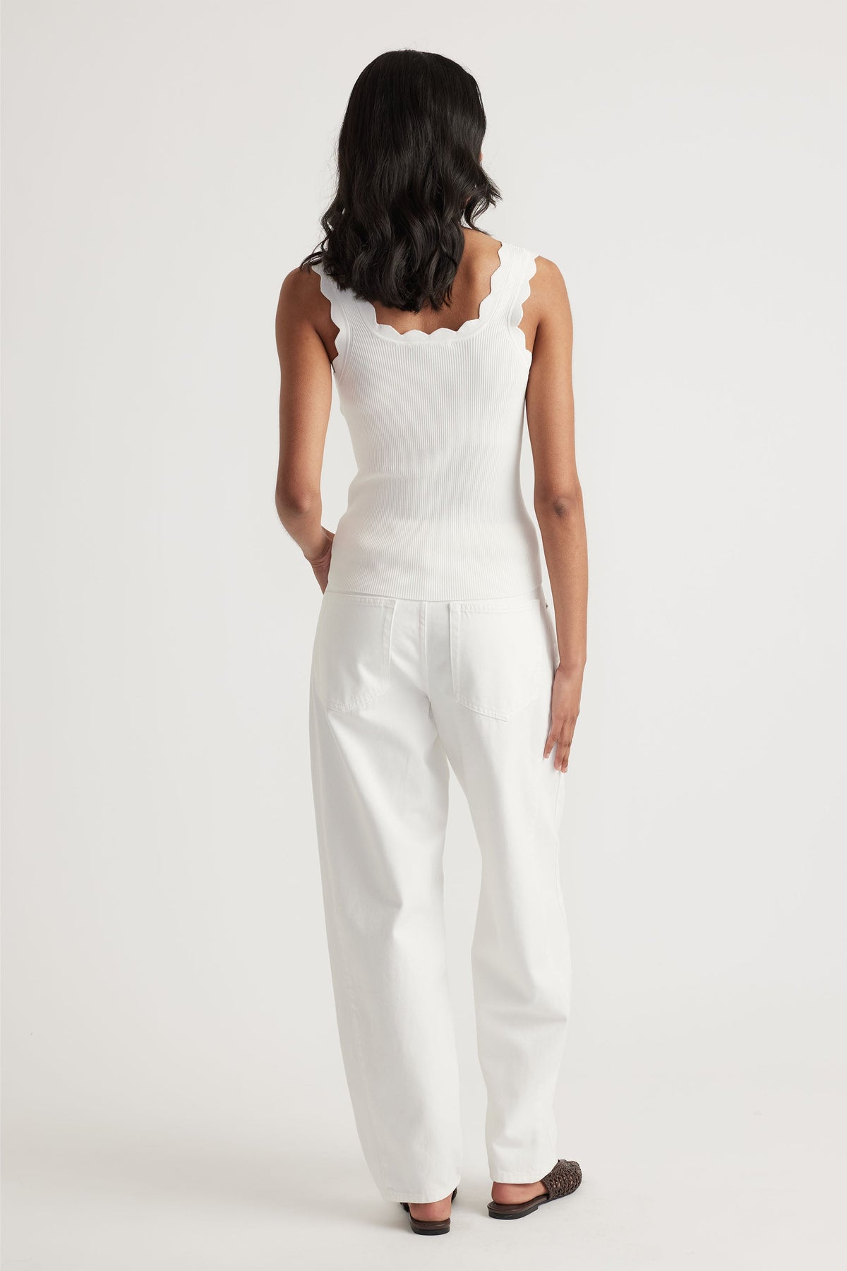 Woman wearing an ivory scallop edge tank with wide straps paired with loose white pants in a minimalist studio setting.