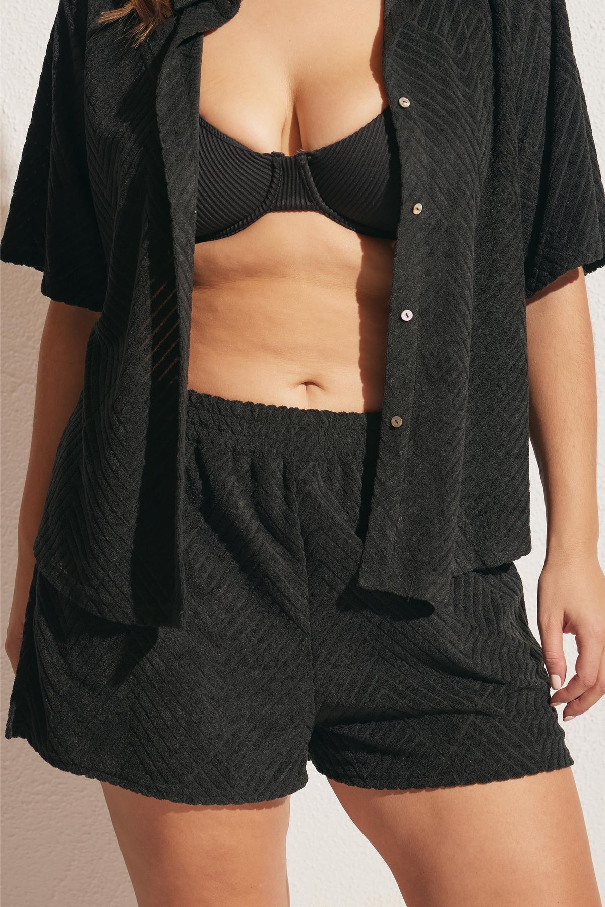 Woman wearing textured black lounge shorts with an open short-sleeve shirt and black ribbed bralette detailing