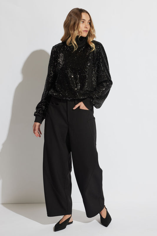 Woman wearing a black sequin turtleneck top paired with wide-leg black pants and pointed slingback shoes, standing.
