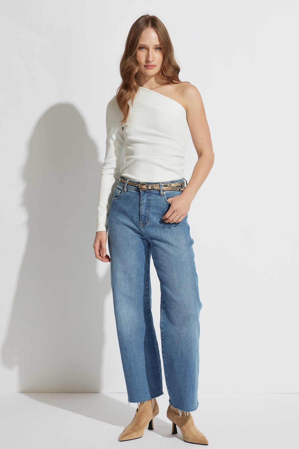 Woman wearing an ivory one-shoulder top paired with high-waisted straight-leg blue jeans and tan ankle boots.