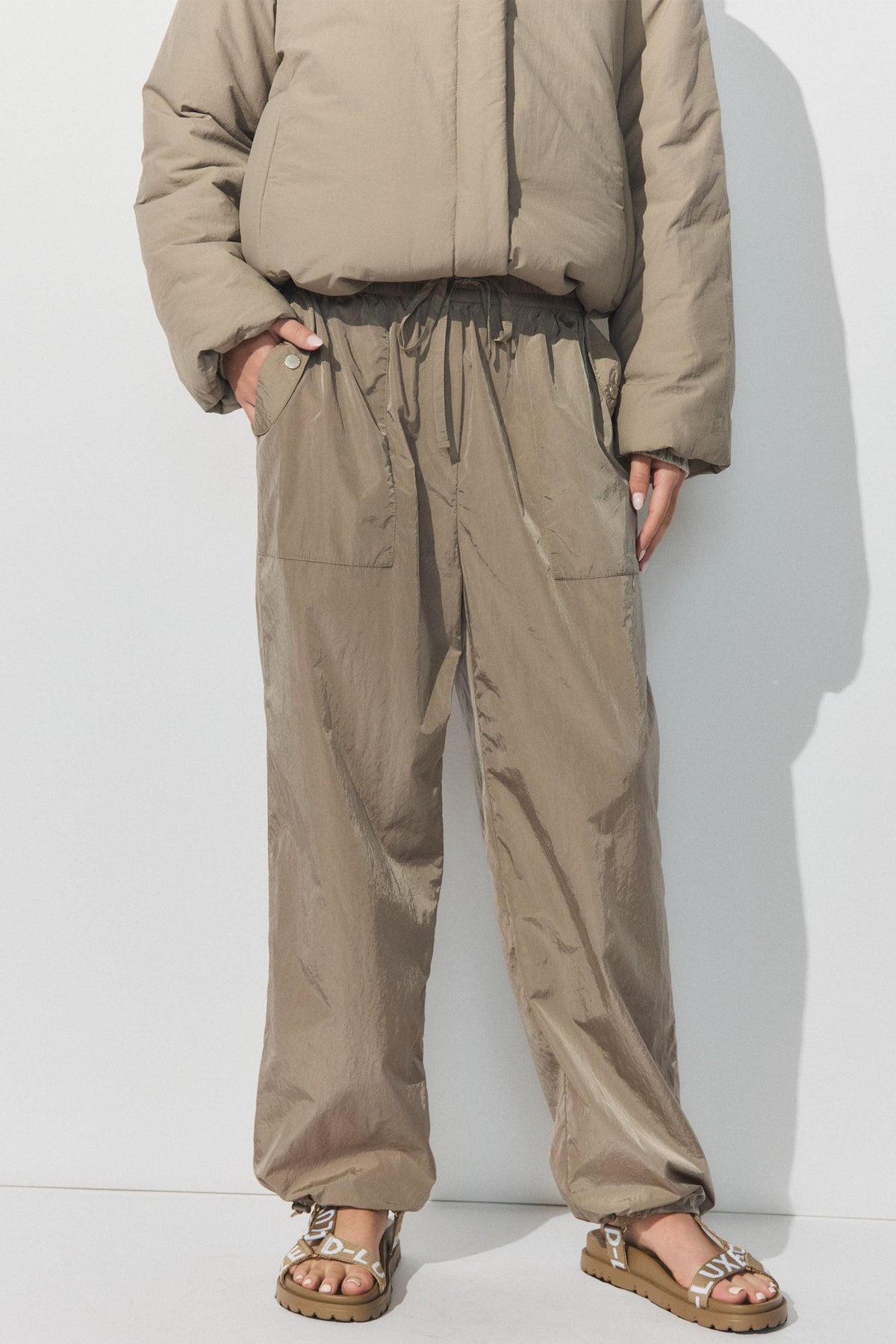 Person wearing loose fit gravel-colored parachute joggers with elastic cuffs and matching puffer jacket, paired with branded sandals.