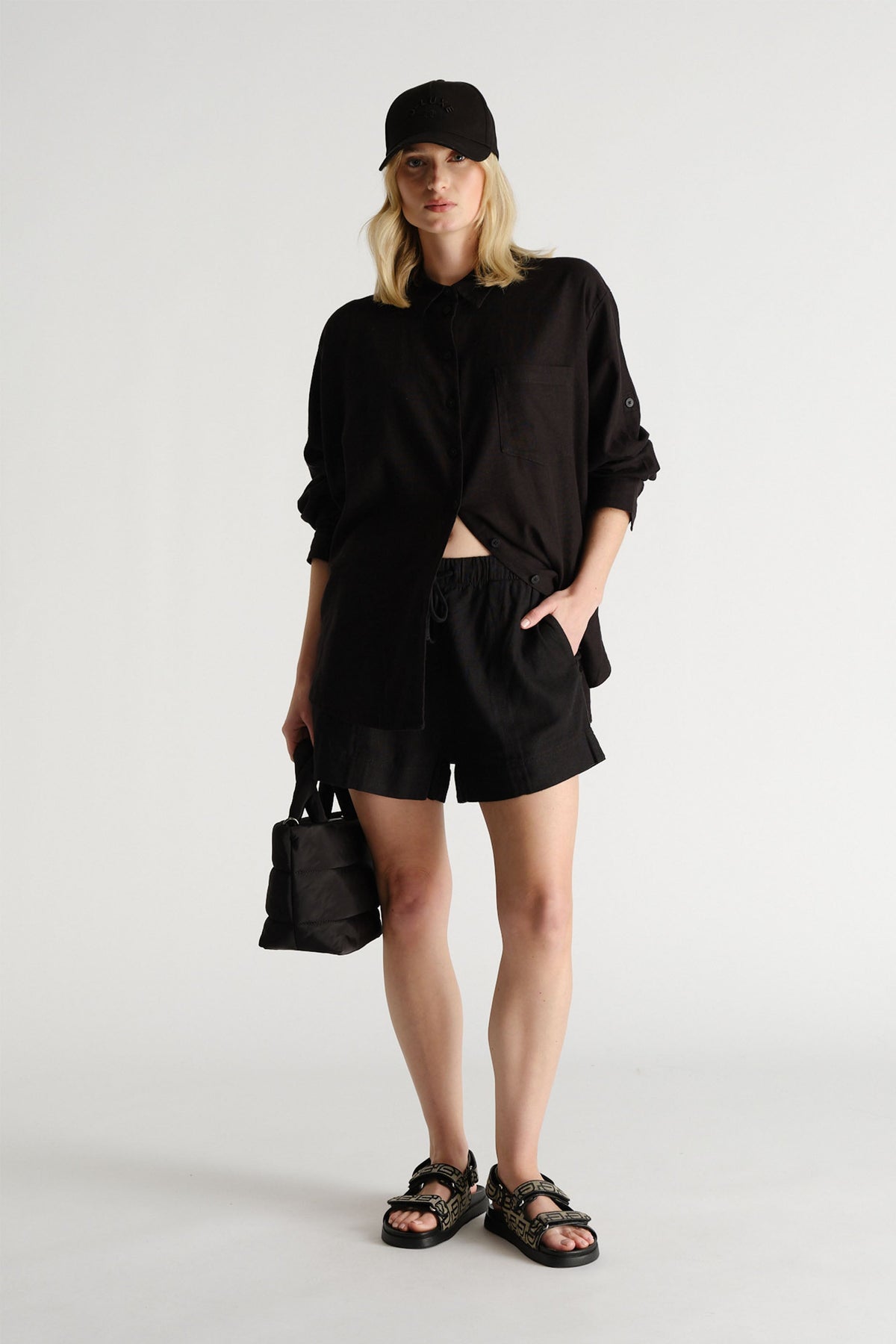 Portrait of a model wearing black D-Luxe Linen Blend Short with matching top, cap, and sandals in gallery position 1.
