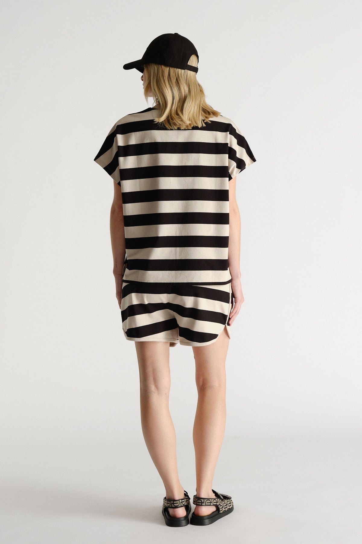 Woman wearing a black and shell color bold striped short set with a black cap and patterned sandals, shown from behind.