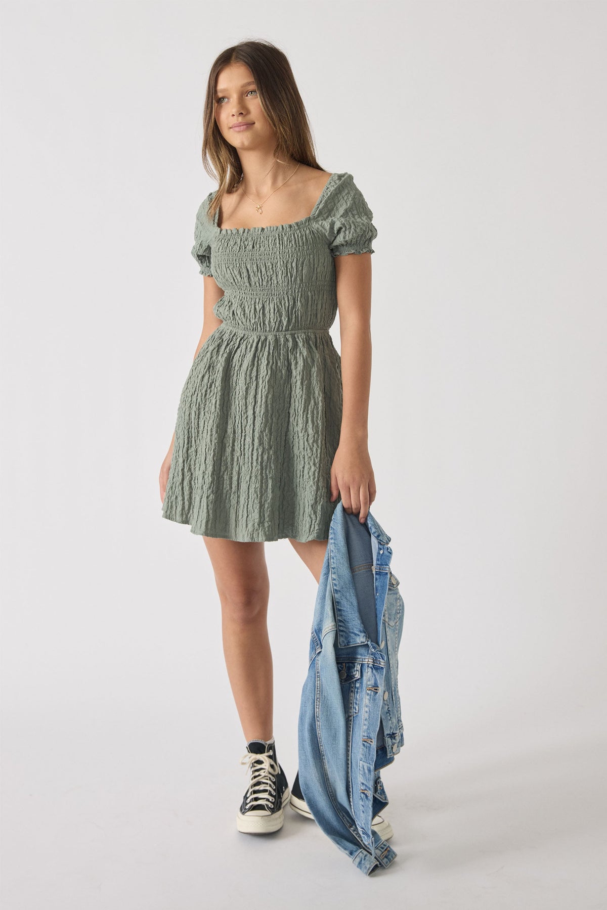 Girl wearing a textured sage green short-sleeve mini dress holding a light wash denim jacket with sneakers on white background