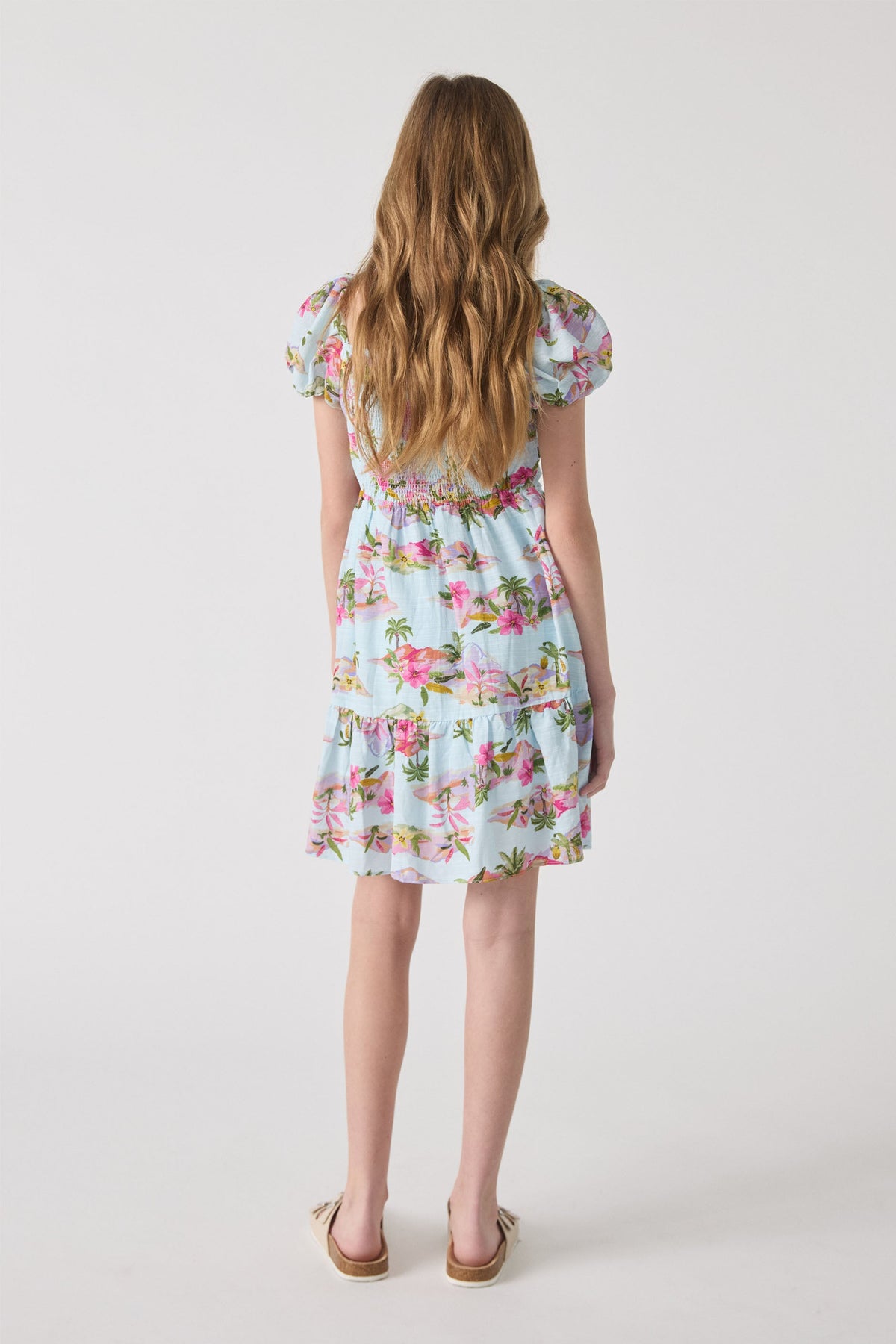 Girl wearing a light blue mini dress with pink floral and palm prints, featuring puff sleeves and a tiered hem.