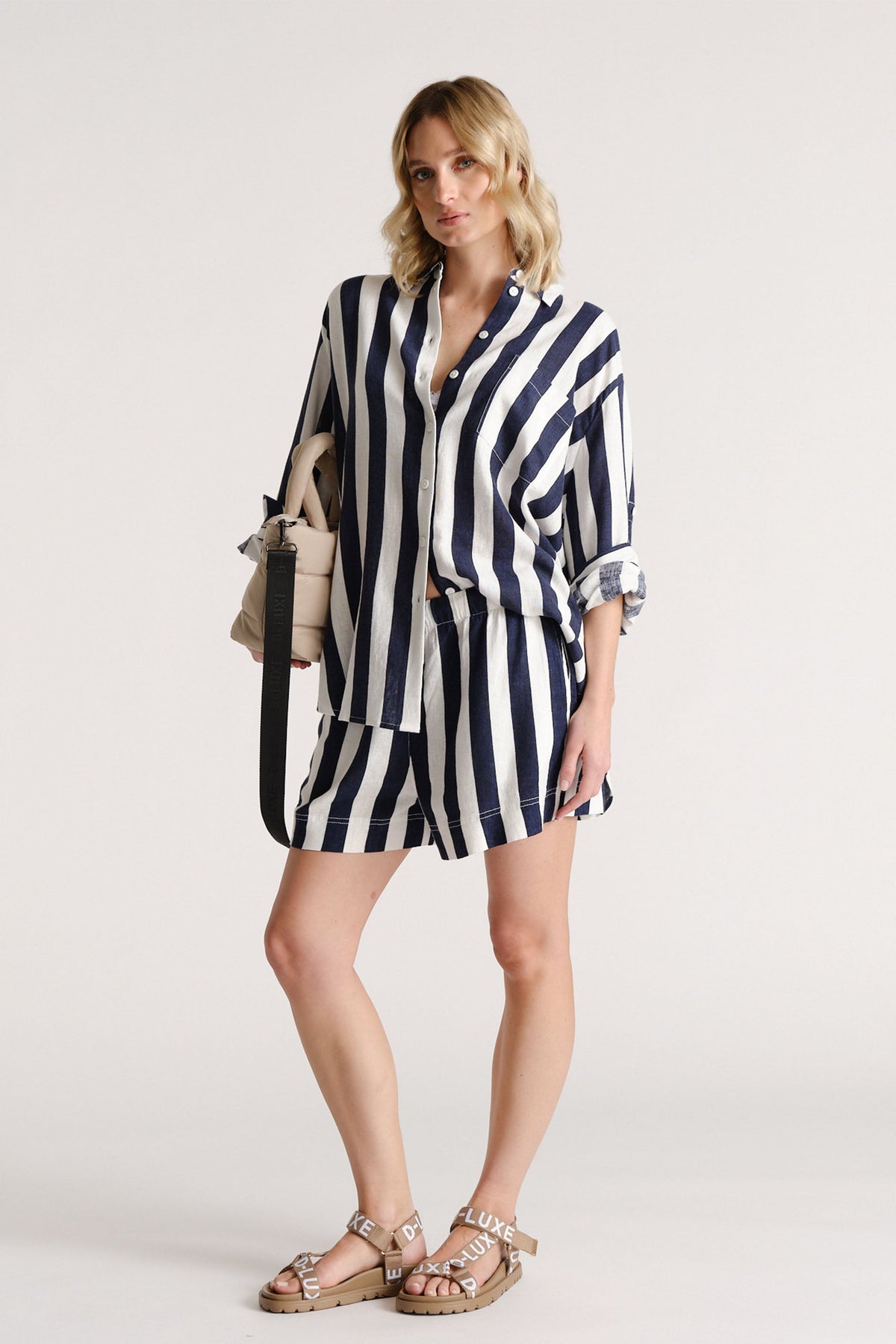 Woman wearing navy and white striped linen blend shorts with matching loose shirt and tan sandals, carrying a beige bag