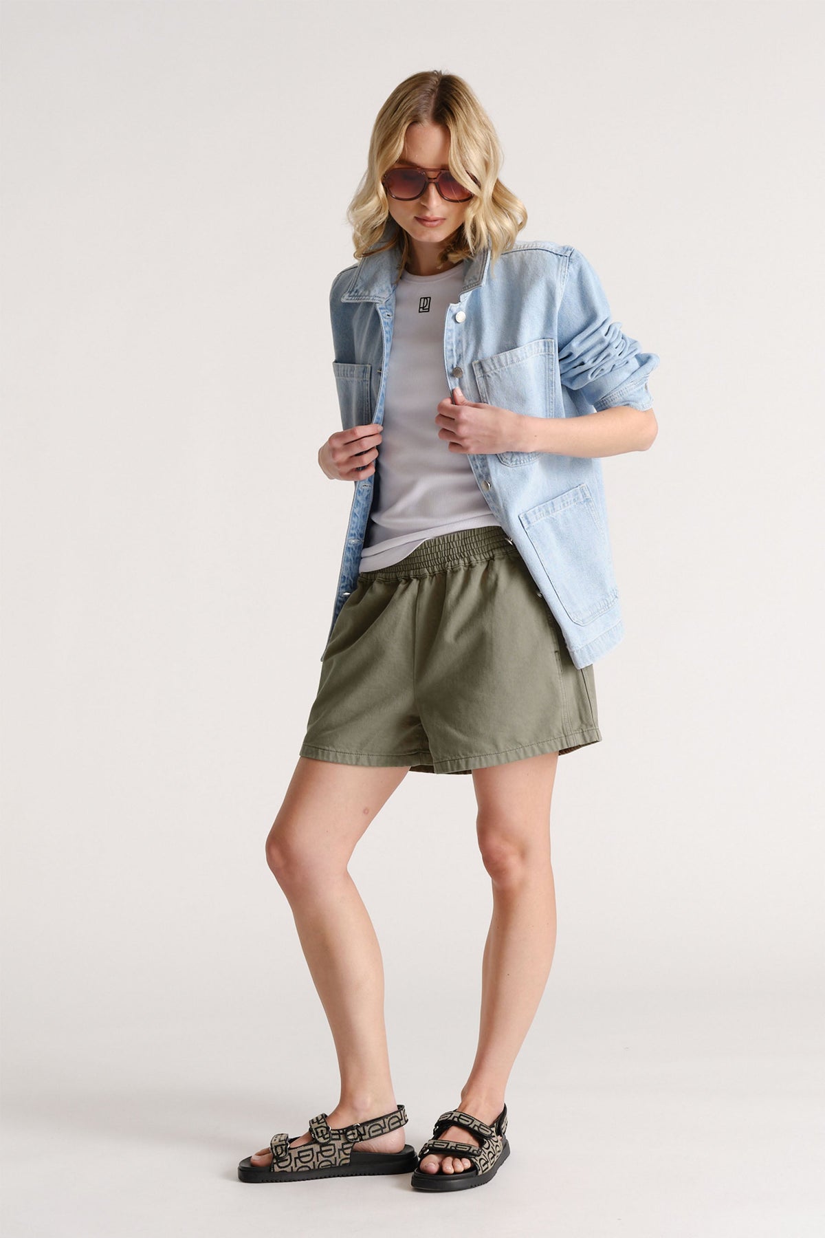Woman wearing olive green pull-on shorts paired with a white tee, light denim jacket, and patterned sandals.