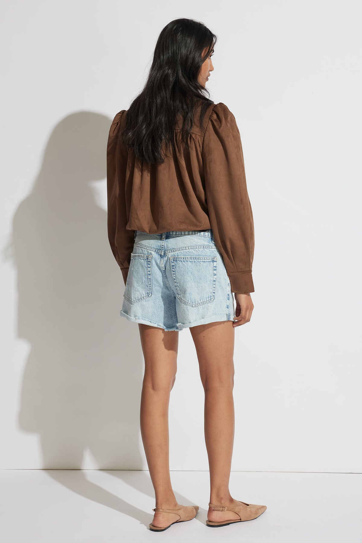 Woman seen from behind wearing light wash rolled cuff denim shorts and a loose brown long sleeve top with puffed shoulders