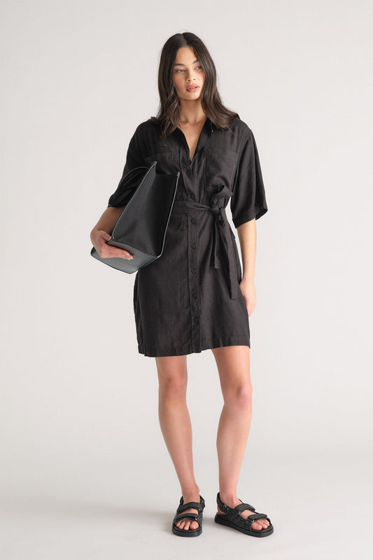 Woman wearing a black linen blend short dress with a waist tie, styled with black sandals and a large black tote bag.
