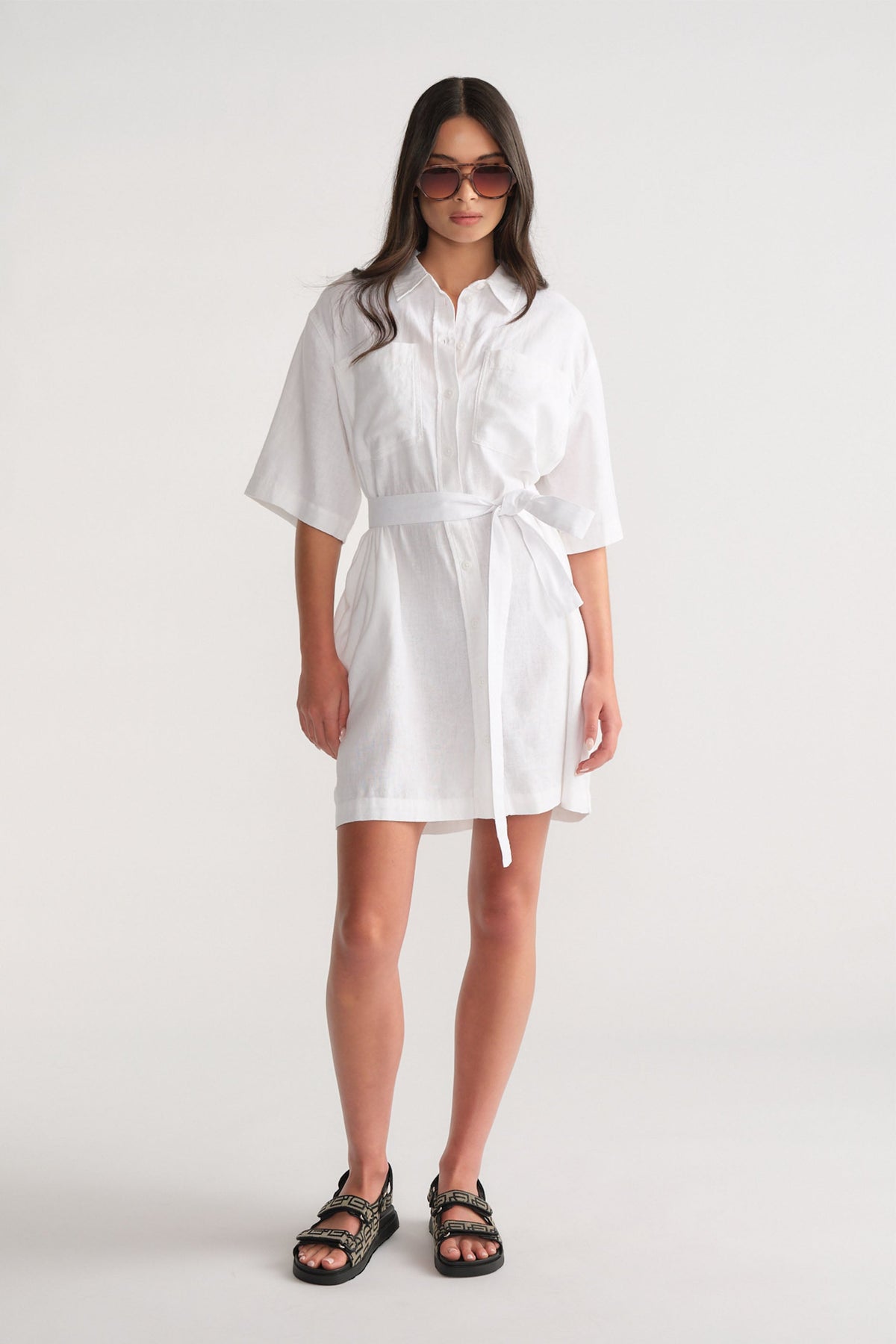 Woman wearing a white linen blend short dress with a self-tie waist, styled with black sandals and oversized sunglasses.