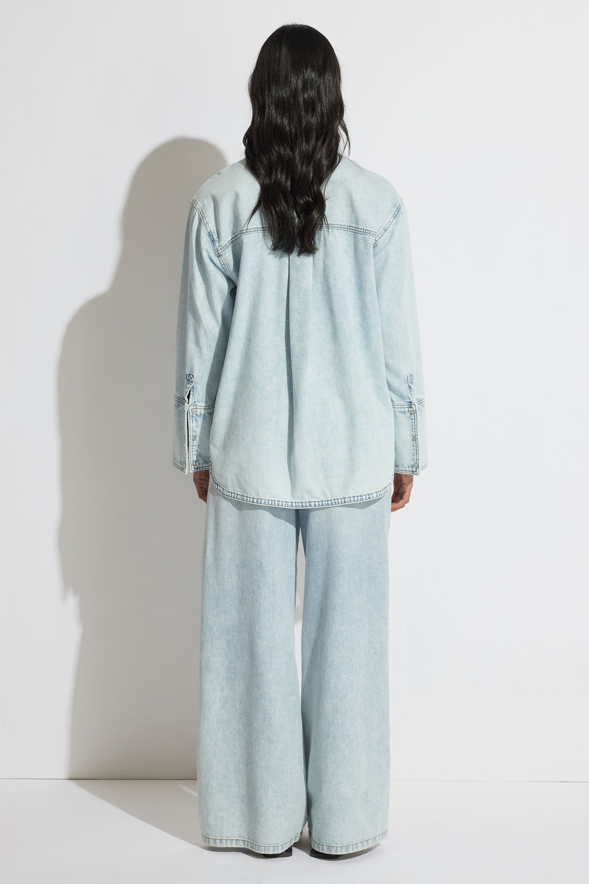 Woman wearing a relaxed fit light blue denim shirt and wide-leg denim pants, shown from the back against a plain wall.