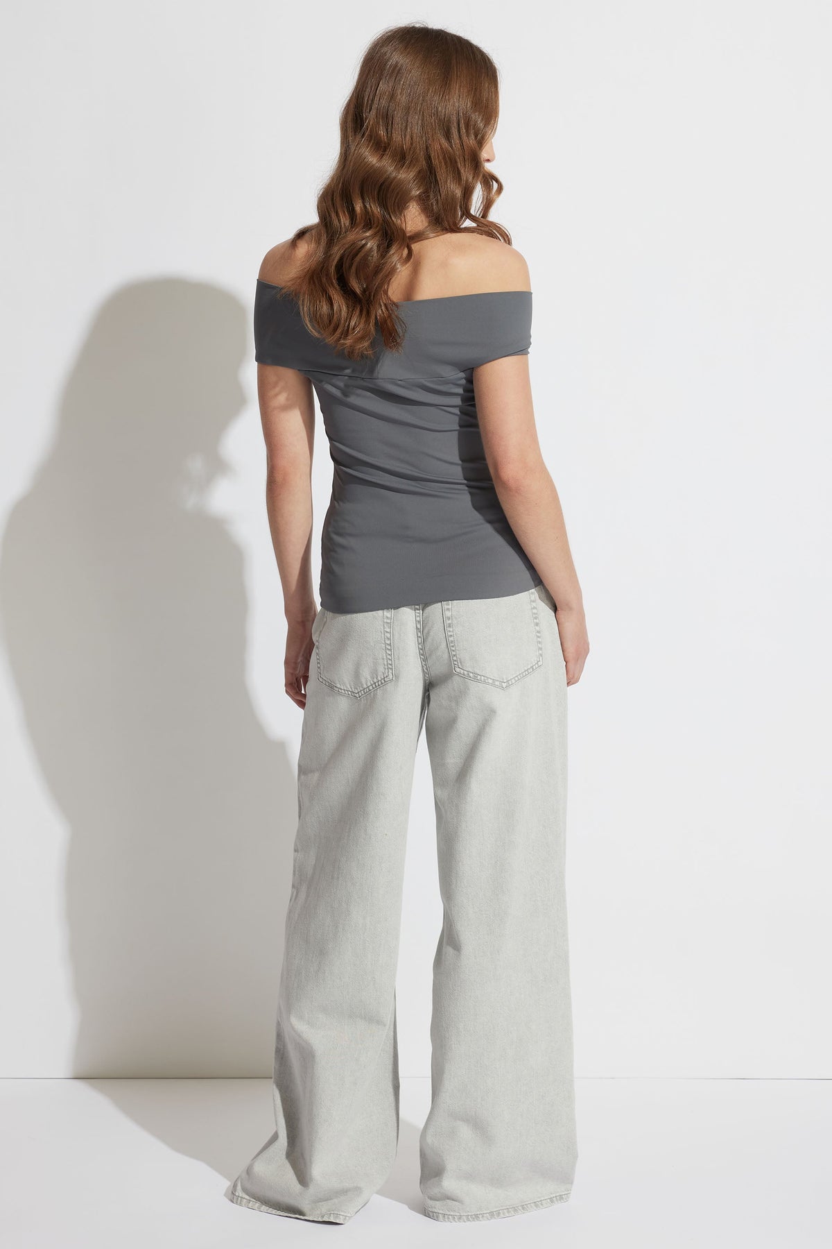 Woman wearing a petrol off-shoulder twist top paired with relaxed-fit, light-wash wide-leg jeans, shown from behind.