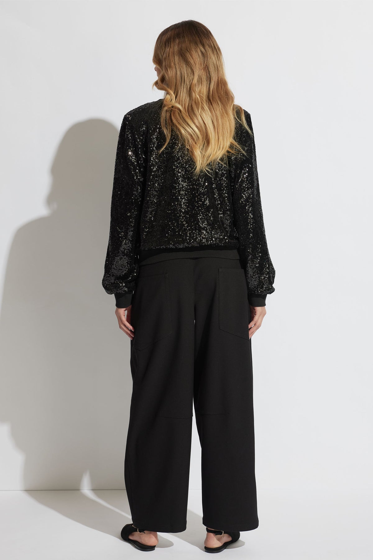 Woman with loose wavy hair wearing a black sequin sweatshirt and wide-leg black pants with flats, seen from behind.