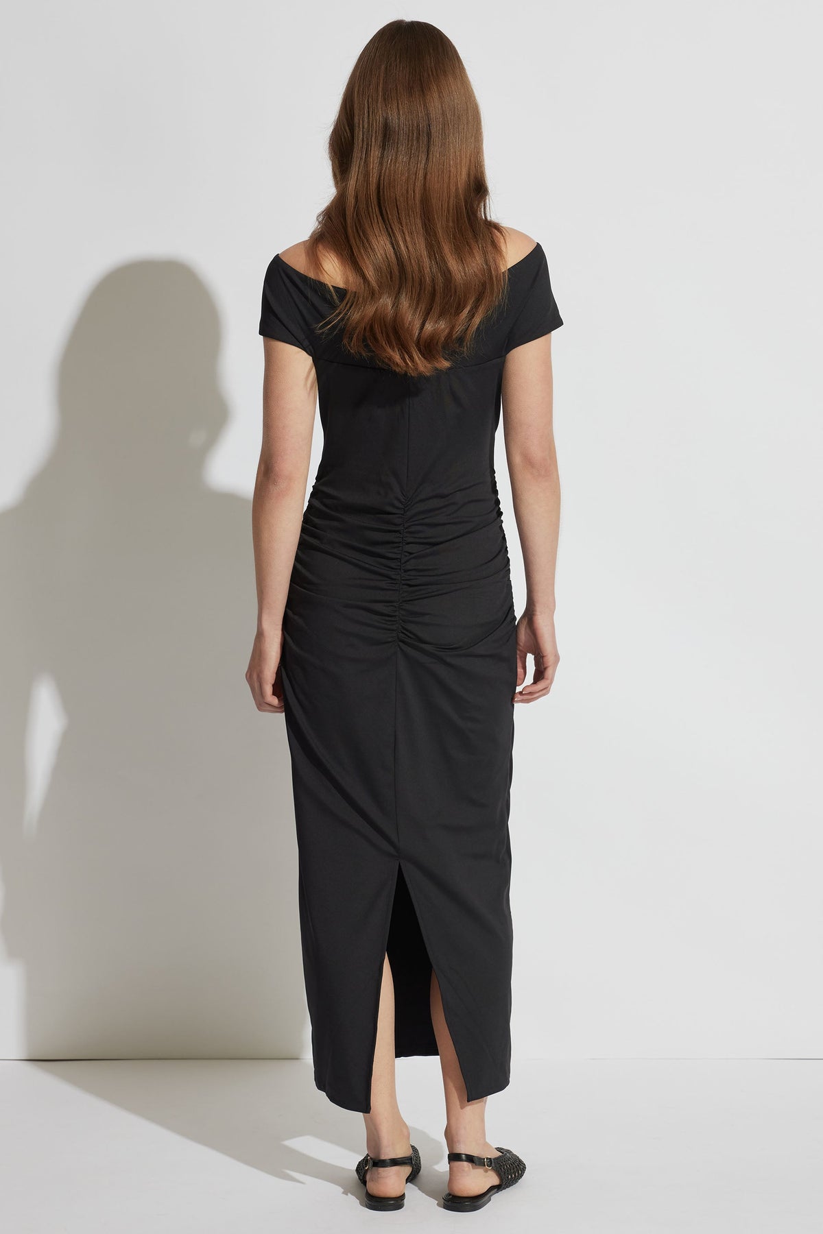 Woman wearing a black off-shoulder midi dress with ruched detailing down the back and a subtle back slit.