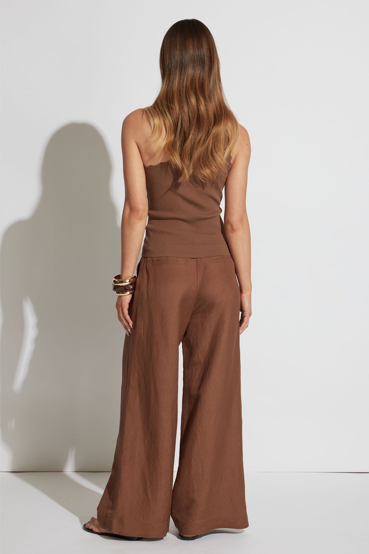 Woman wearing a brown one-shoulder tank paired with wide-leg pants and stacked bangles, shown from behind.