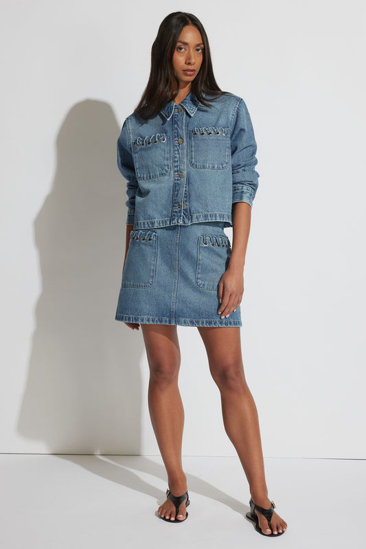 Woman wearing a bright blue denim jacket and matching skirt with unique metal ring details styled with black sandals