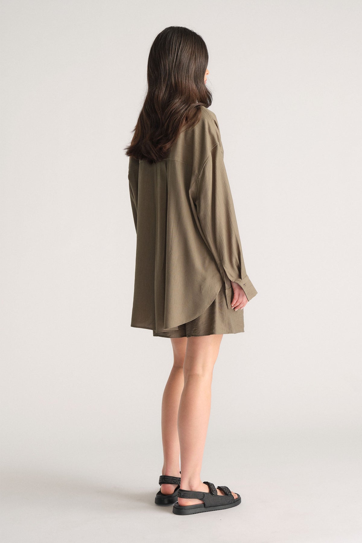 Girl wearing an oversized olive green textured shirt paired with matching shorts and black sandals, viewed from behind.