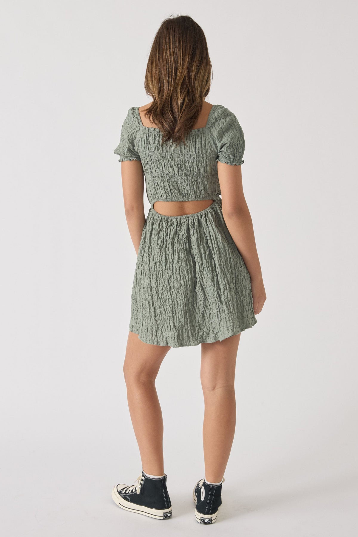 Girl wearing a textured sage green mini dress with puffed sleeves and a cutout back, paired with black high-top sneakers