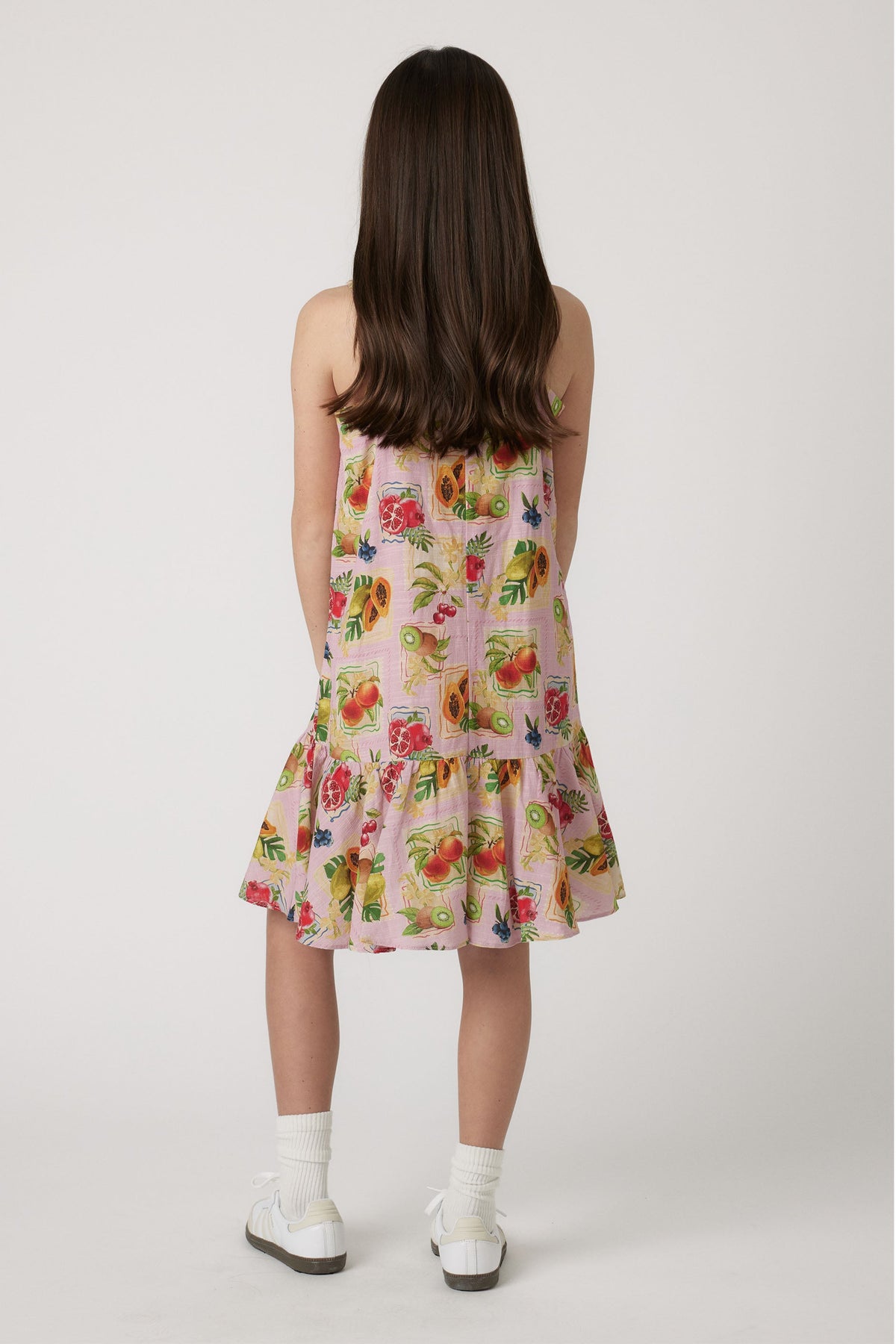 Girl wearing a pink strappy dress with colorful fruit print, paired with white socks and sneakers seen from behind