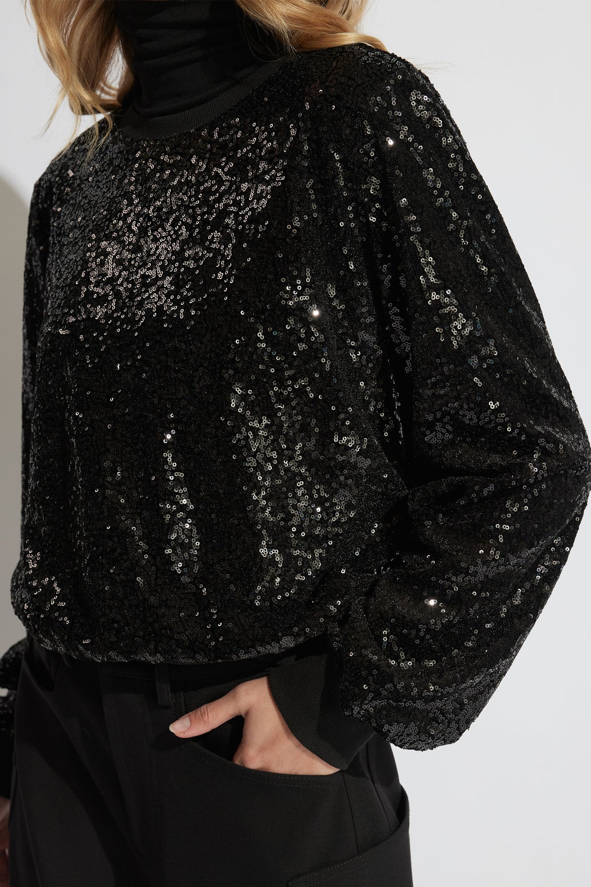 Close-up of woman wearing a black sequin sweat with puffed sleeves and a ribbed collar, styled with black pants