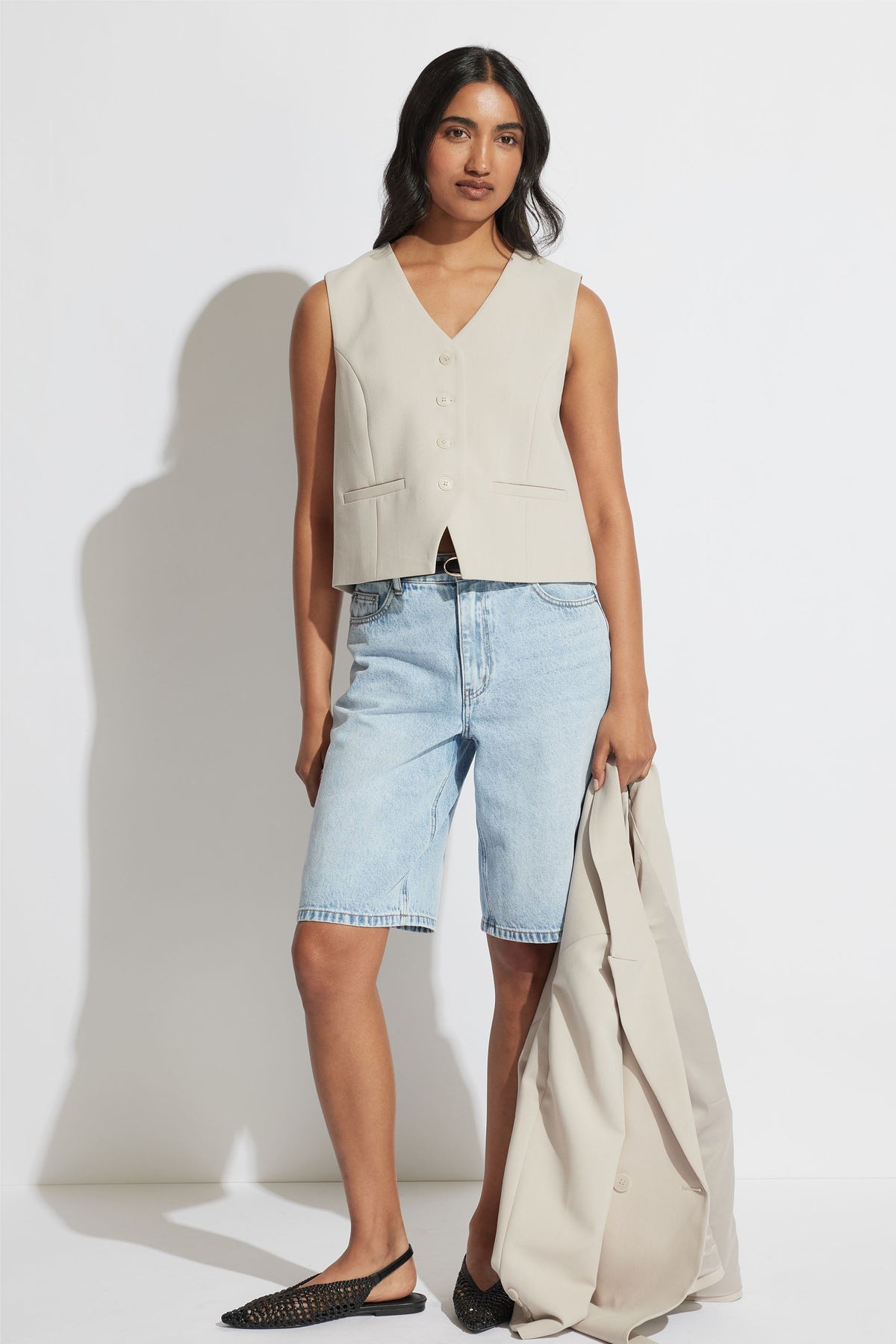 Woman wearing a tailored cream vest with light blue denim long shorts and black woven flats, holding a matching jacket.