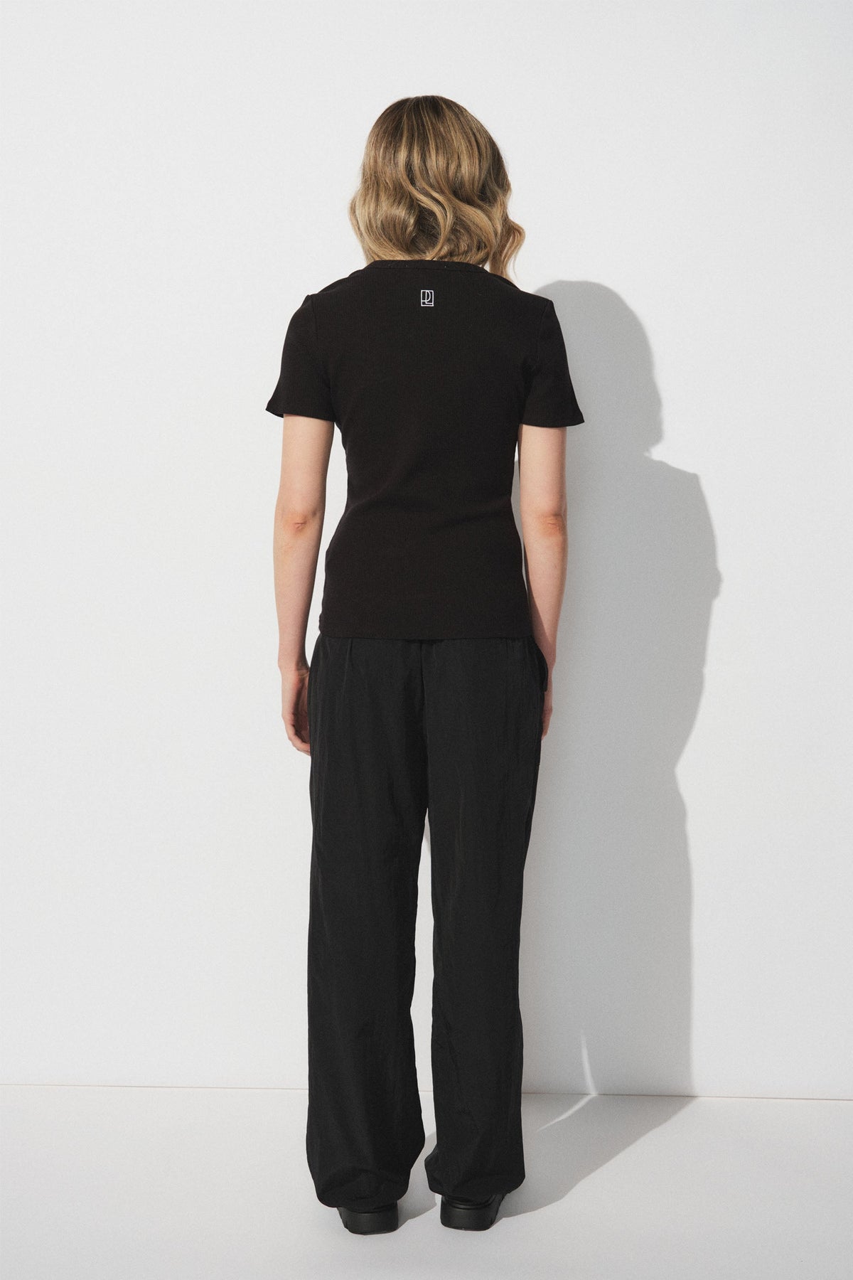 Woman with wavy blonde hair wearing a fitted black short-sleeve top and relaxed black parachute-style joggers, back view.
