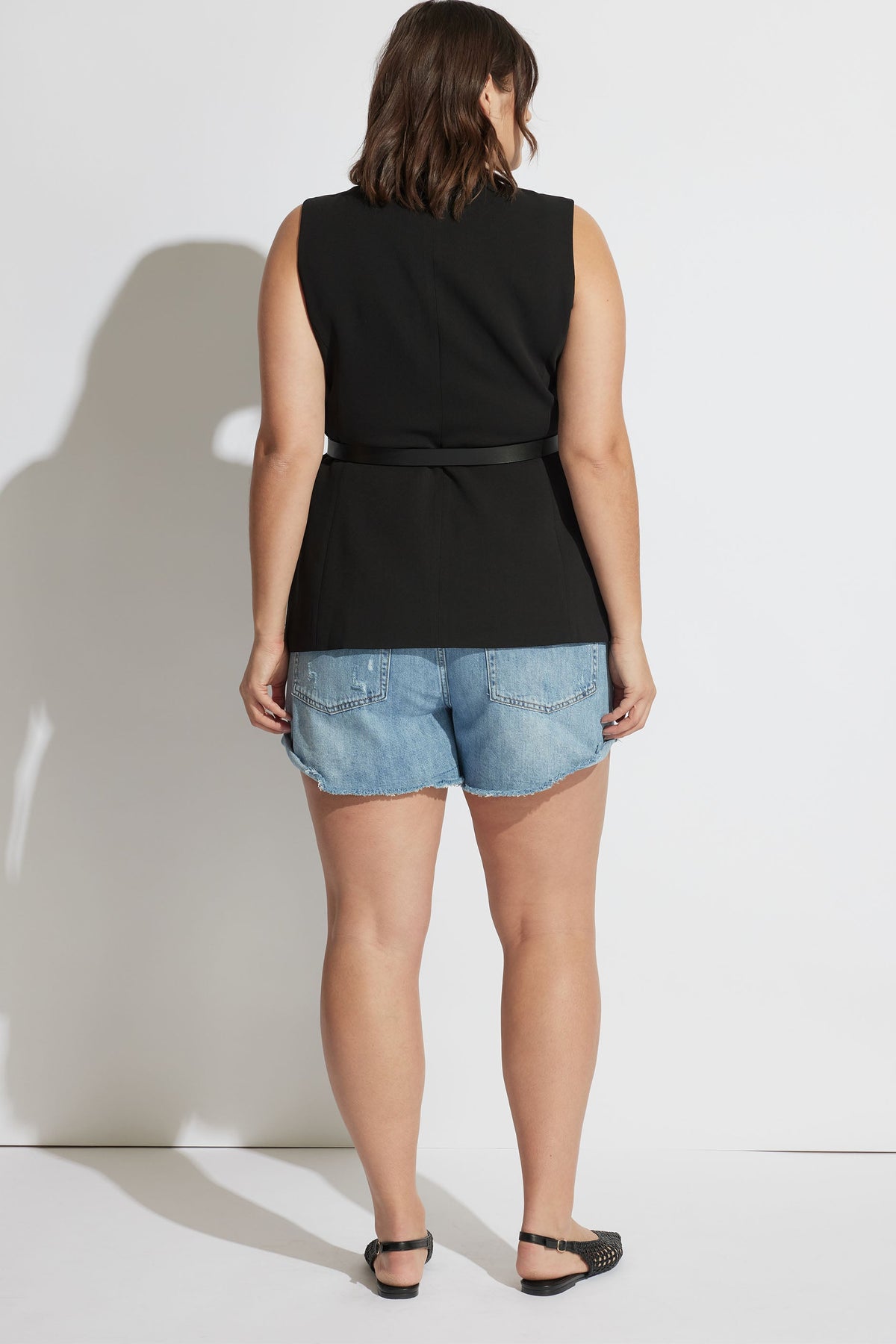 Woman wearing light blue rolled cuff denim shorts paired with a sleeveless black belted top and black flats.