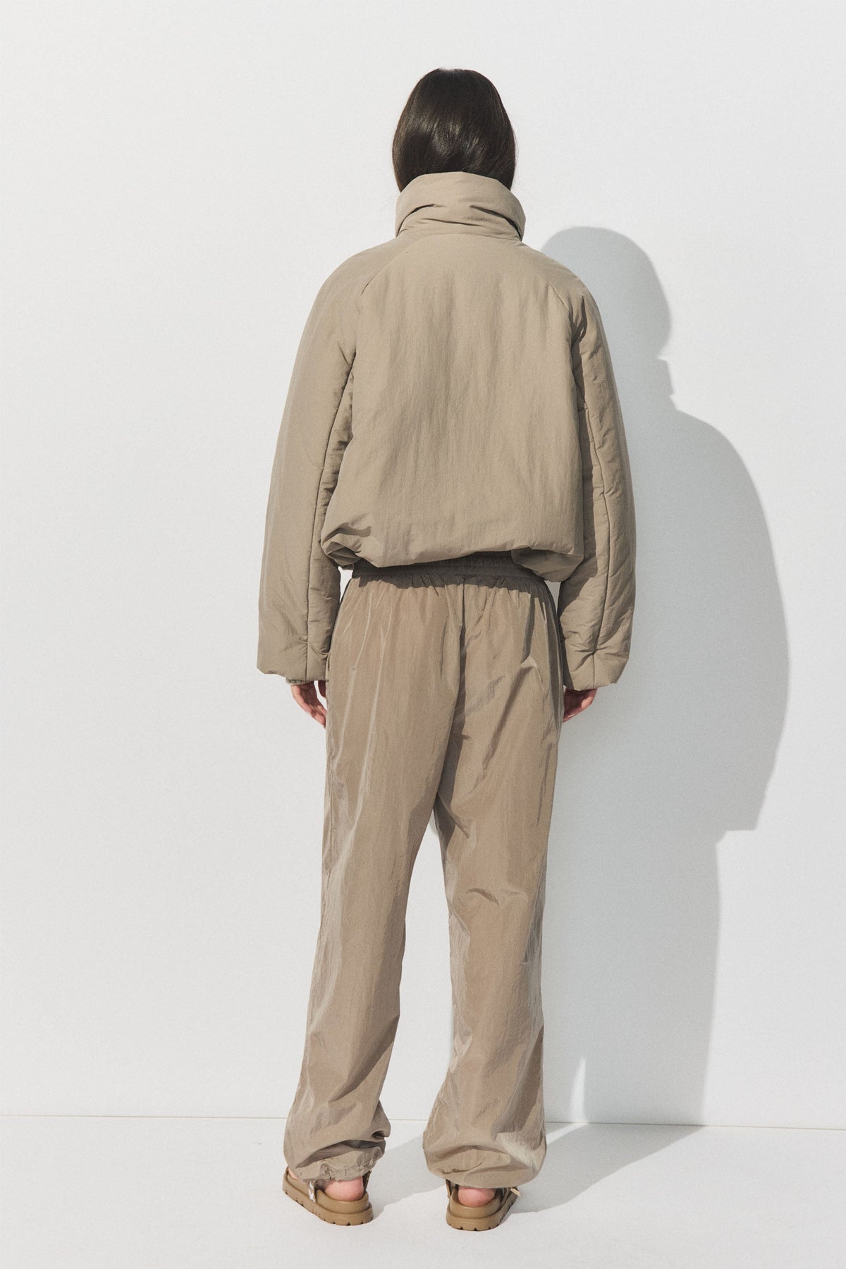 Woman wearing a loose beige padded jacket and relaxed-fit joggers standing against a white wall, back view.