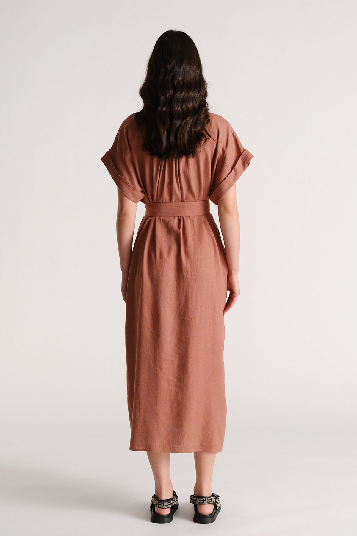 Woman seen from behind wearing a rust-colored linen blend midi dress with cuffed sleeves and a wide belt at the waist