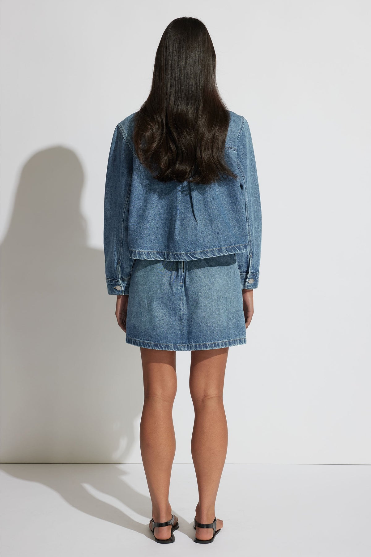 Woman facing away wearing a relaxed fit bright blue denim jacket paired with a matching denim mini skirt and black sandals