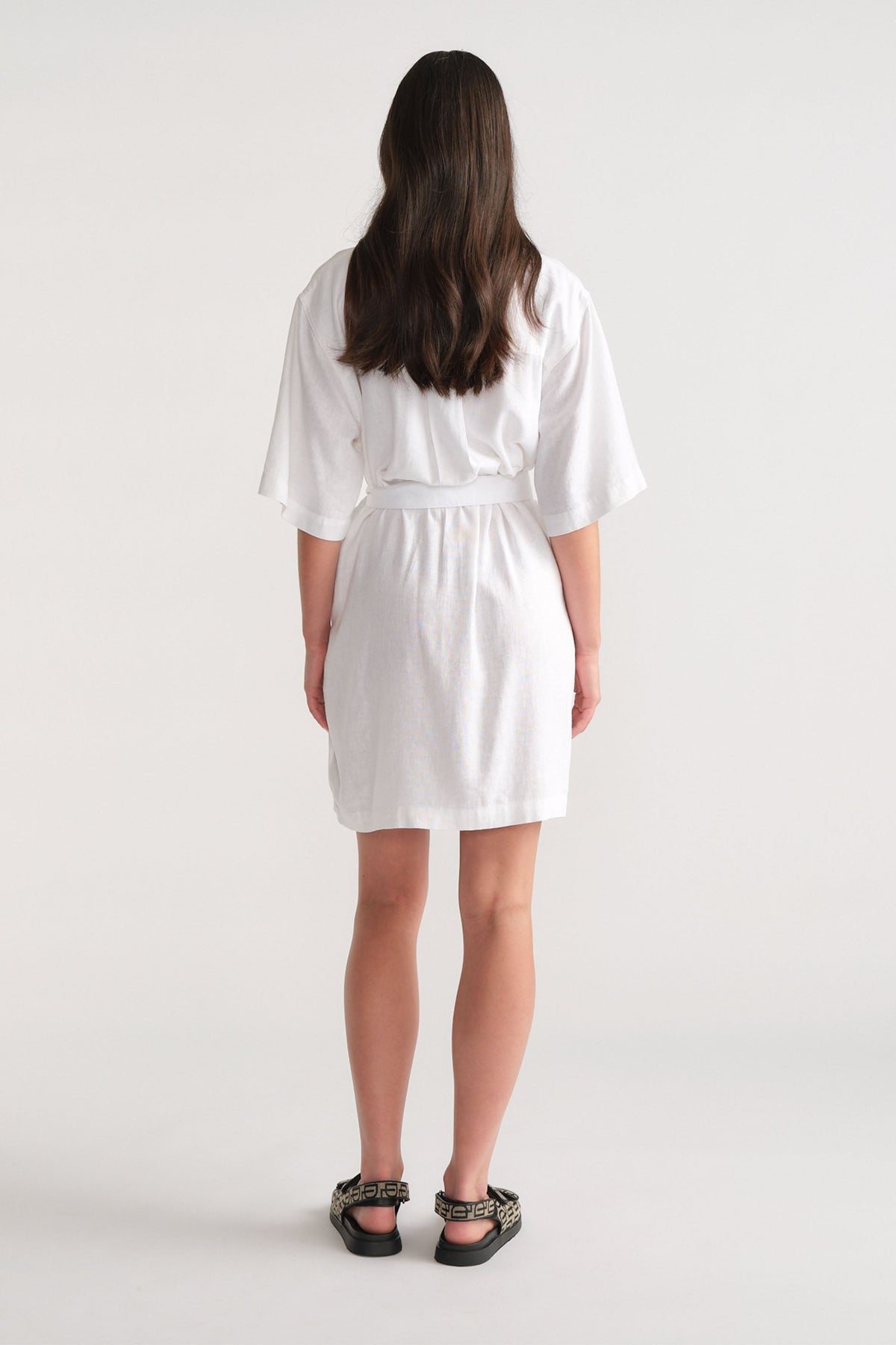 Woman wearing a white short linen blend dress with loose sleeves, styled with patterned black sandals, shown from the back.
