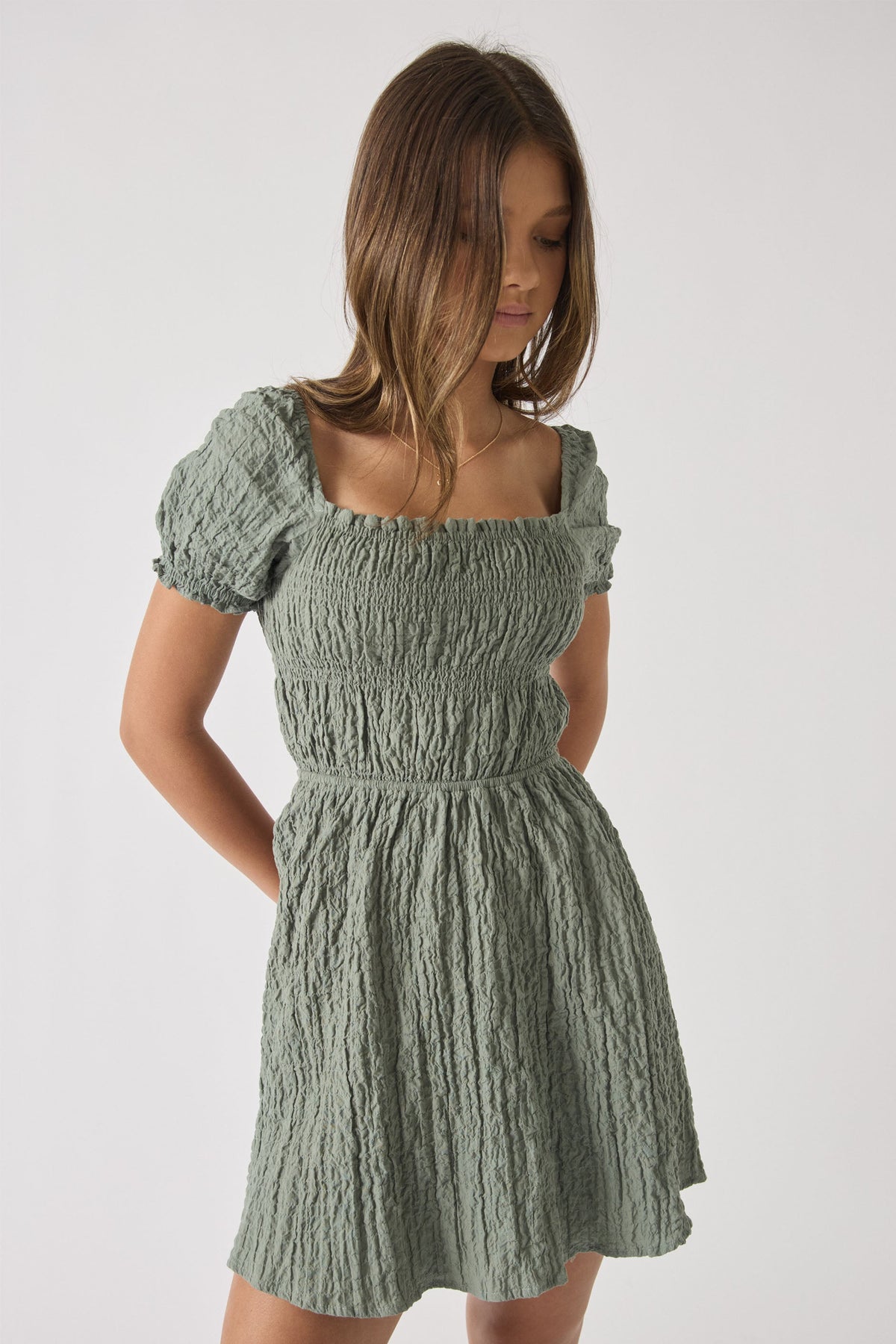 Girl wearing a textured sage green mini dress with puff sleeves and a square neckline, looking downward.