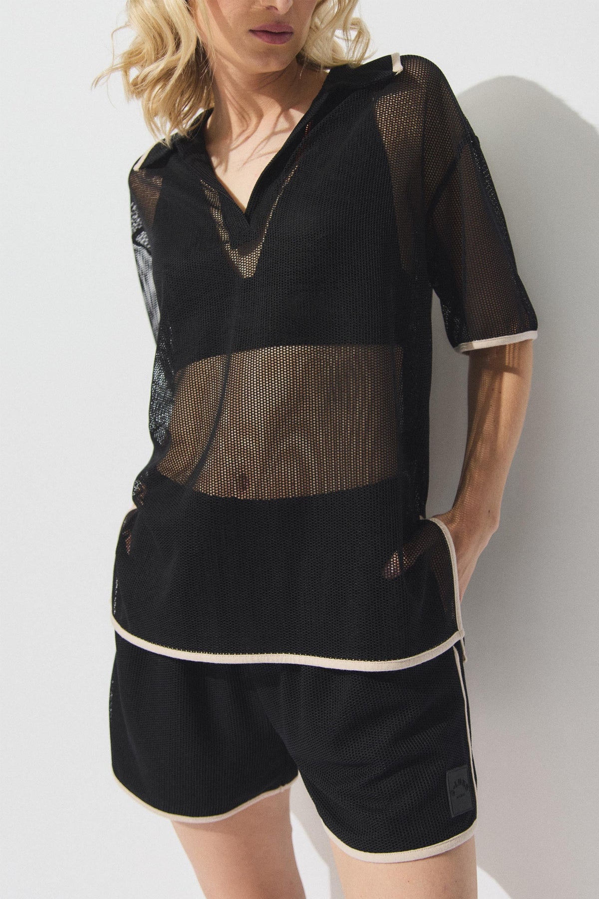 Woman wearing black mesh knit polo top with white trim and matching shorts, posing with hands in pockets against light background