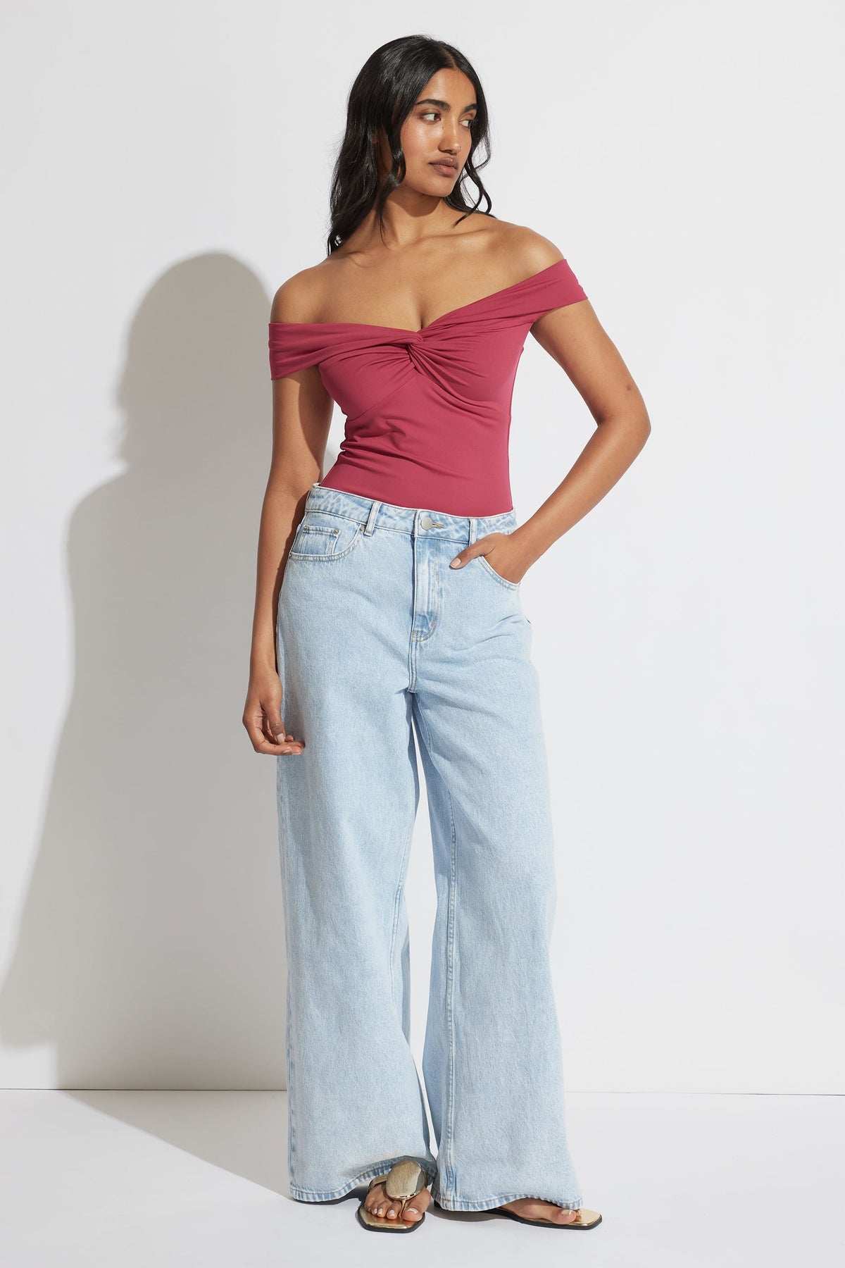 Woman wearing a twisted off-shoulder cerise top paired with light wash wide-leg jeans and slide sandals