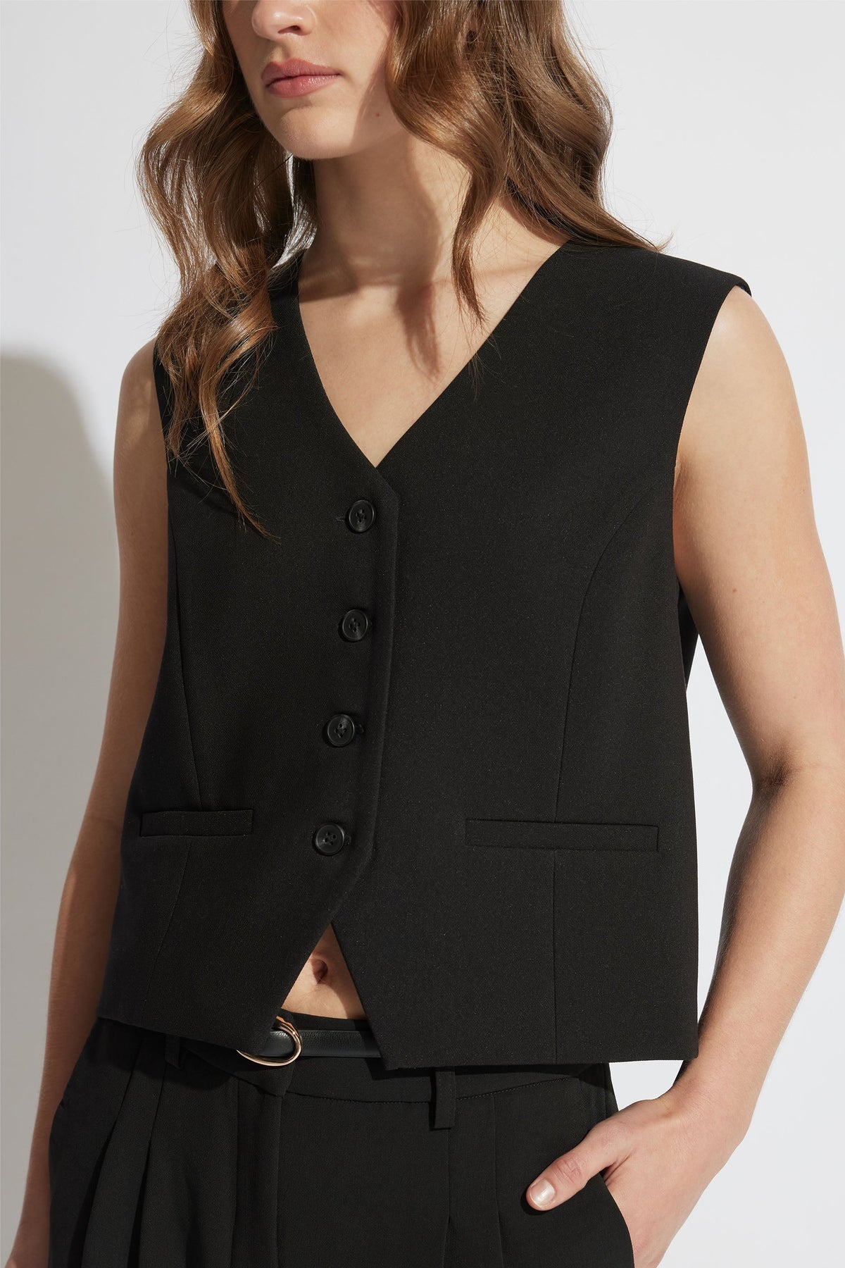 Sleeveless black tailored vest with V-neck and button front, styled with high-waisted black pants and belt.