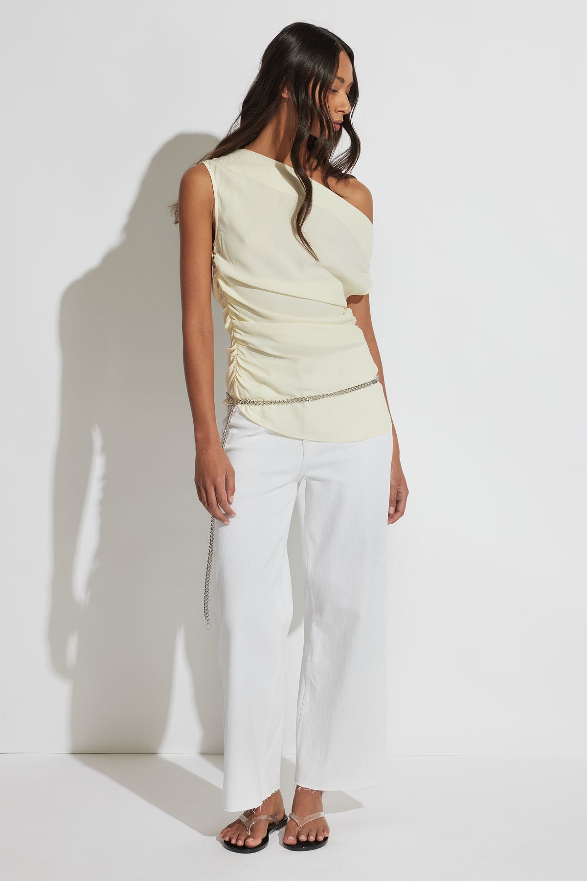 Woman wearing a pale yellow one-shoulder ruched top with white wide-leg pants and sandals, accessorized with a chain belt
