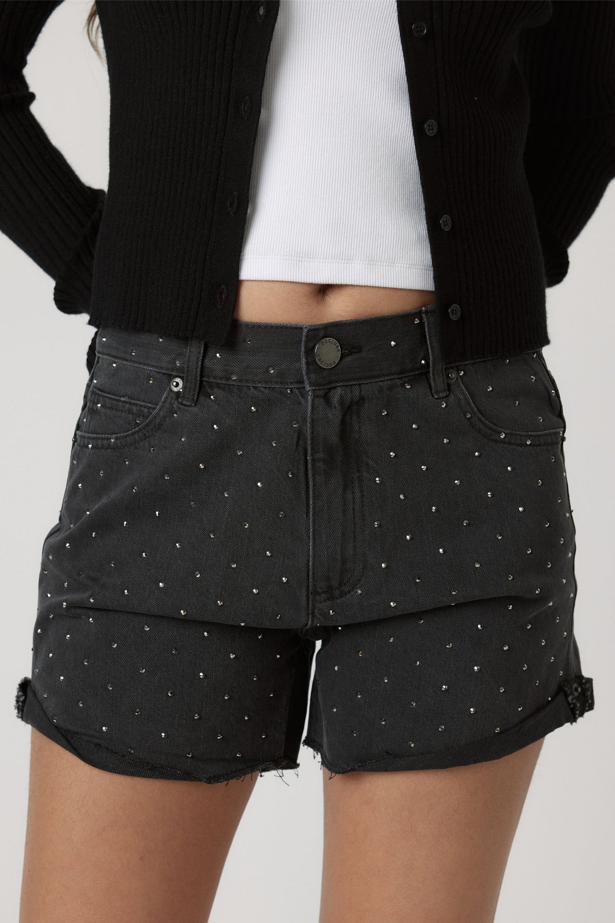 Close-up of black denim shorts embellished with small rhinestones, styled with a cropped white top and black cardigan.