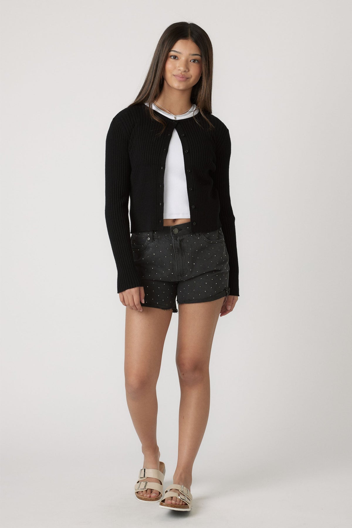 Teen girl wearing black rhinestone-embellished shorts paired with a cropped black ribbed cardigan and white slide sandals