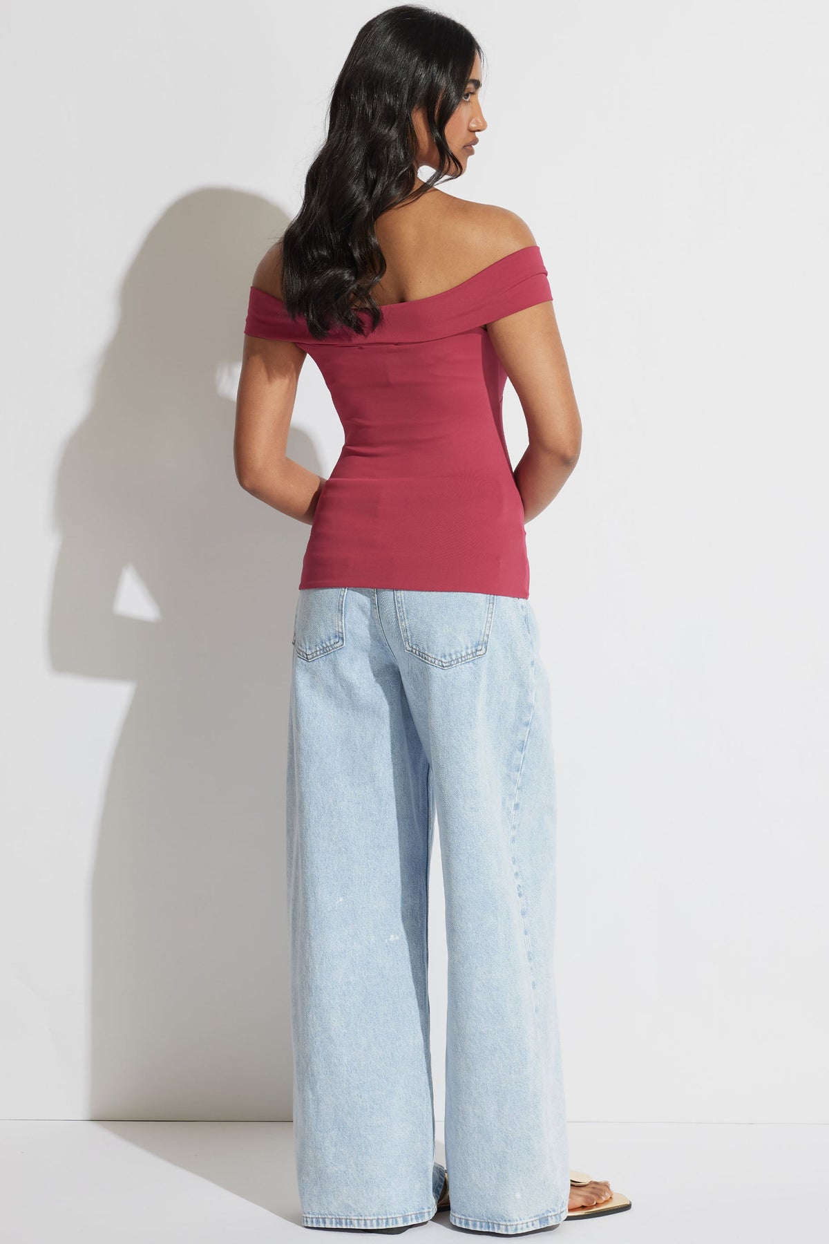 Woman wearing a deep rose off-shoulder twist top paired with light wide-leg denim jeans and sandals, posing sideways.