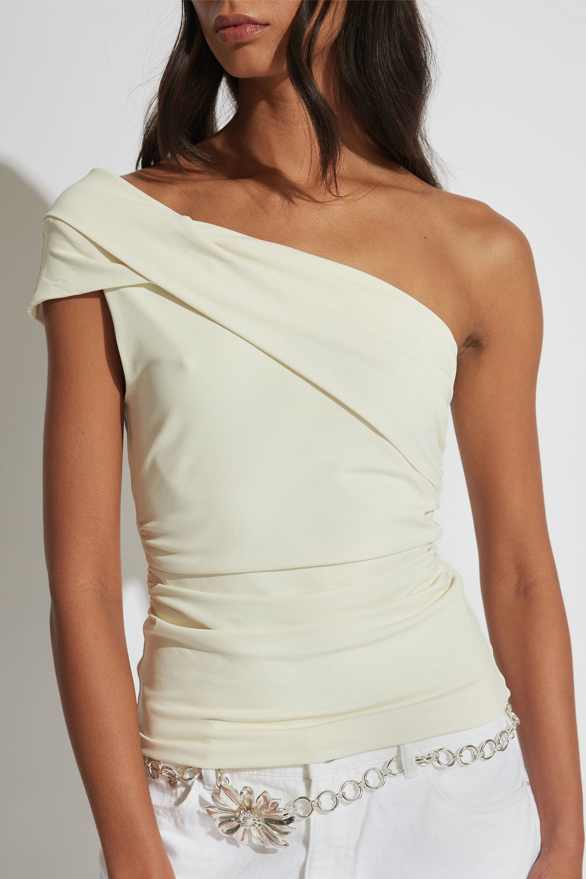 Woman wearing an ivory one-shoulder twist top paired with white pants and a silver floral chain belt.
