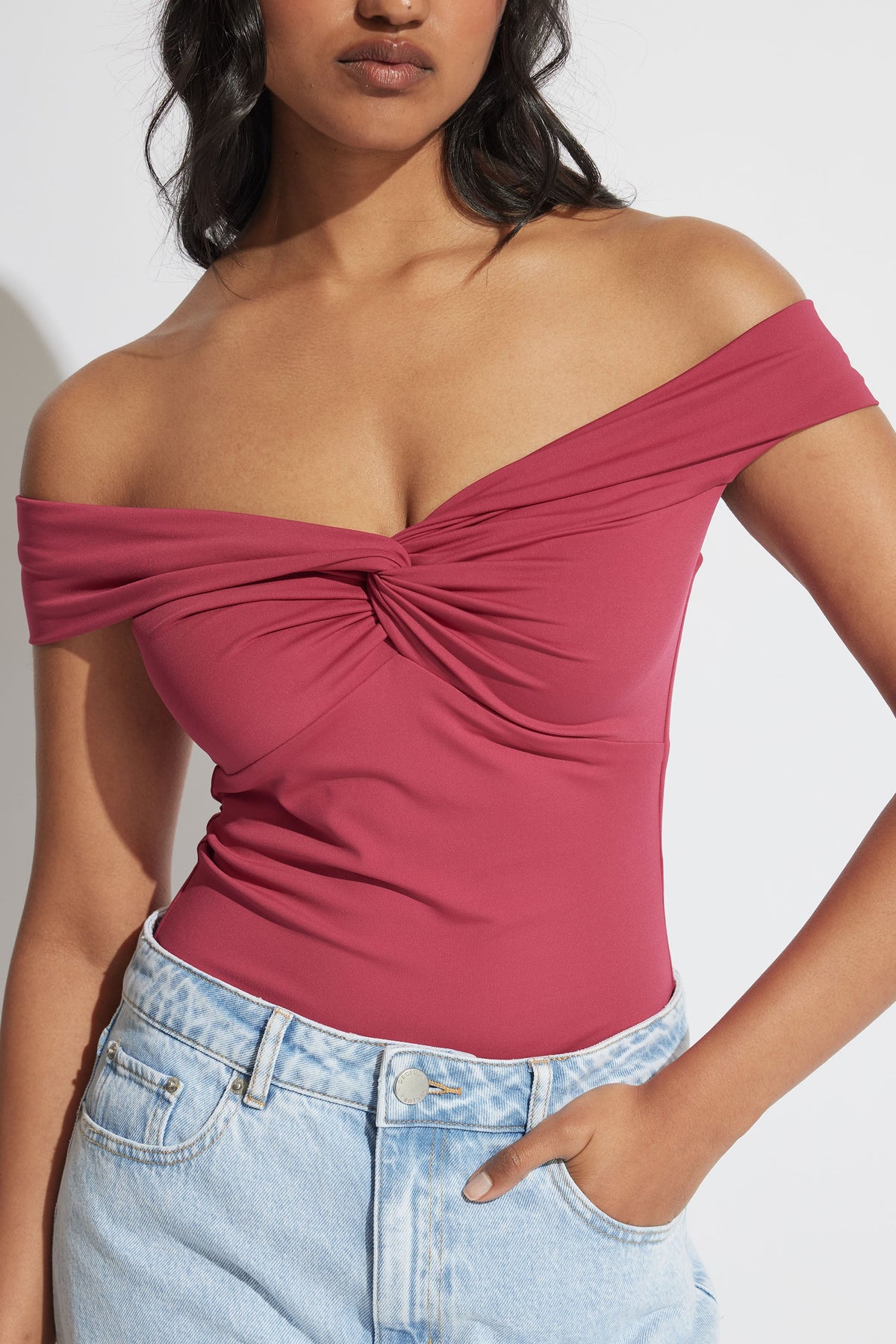 Woman wearing a cerise off-shoulder twist top paired with light wash denim jeans in soft natural light