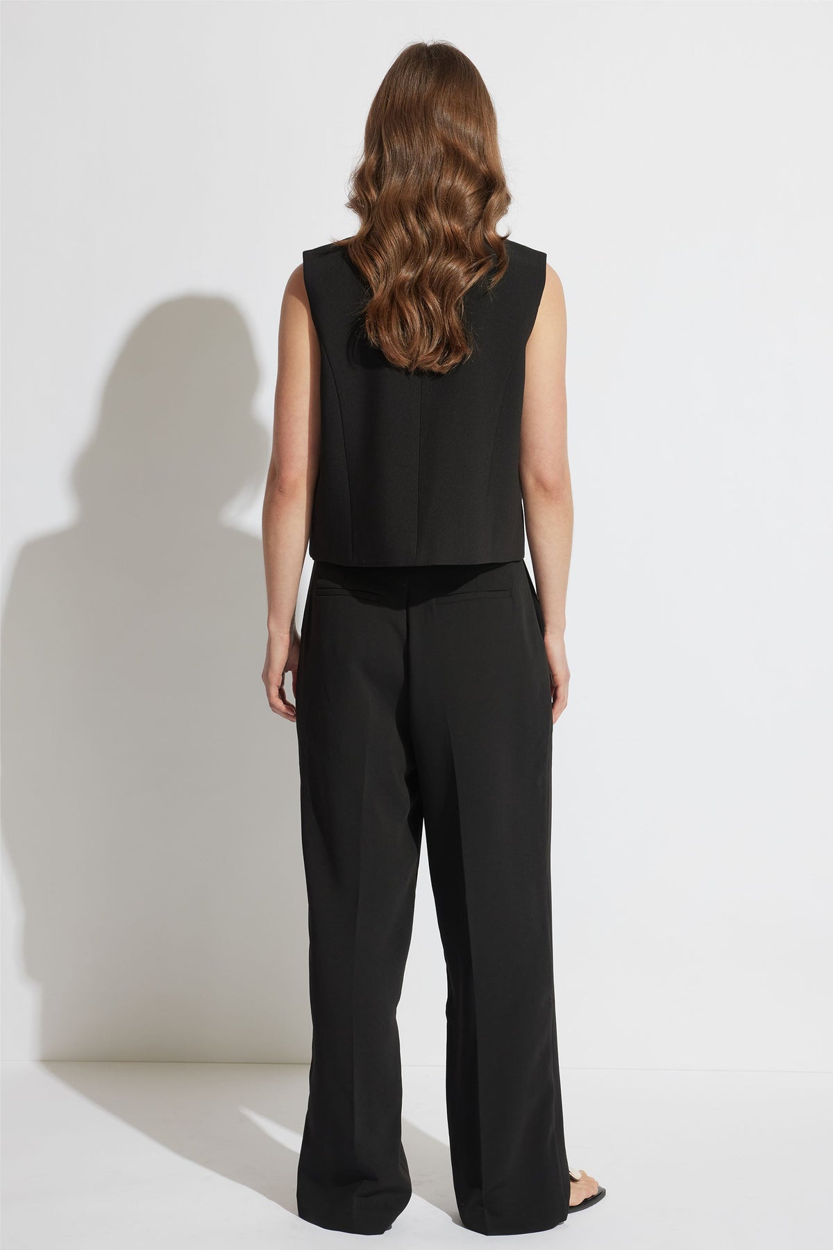 Woman seen from behind wearing a black tailored sleeveless vest paired with loose black trousers and sandals