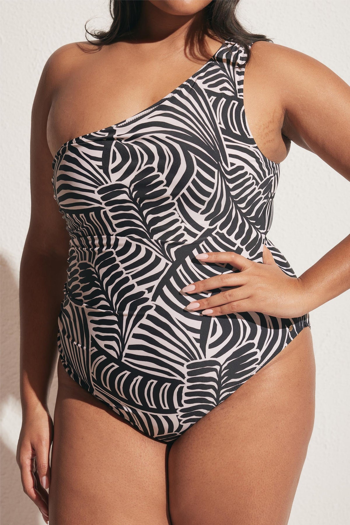 Curvy woman wearing a matte one-shoulder one piece swimsuit with black and pale pink abstract leaf print